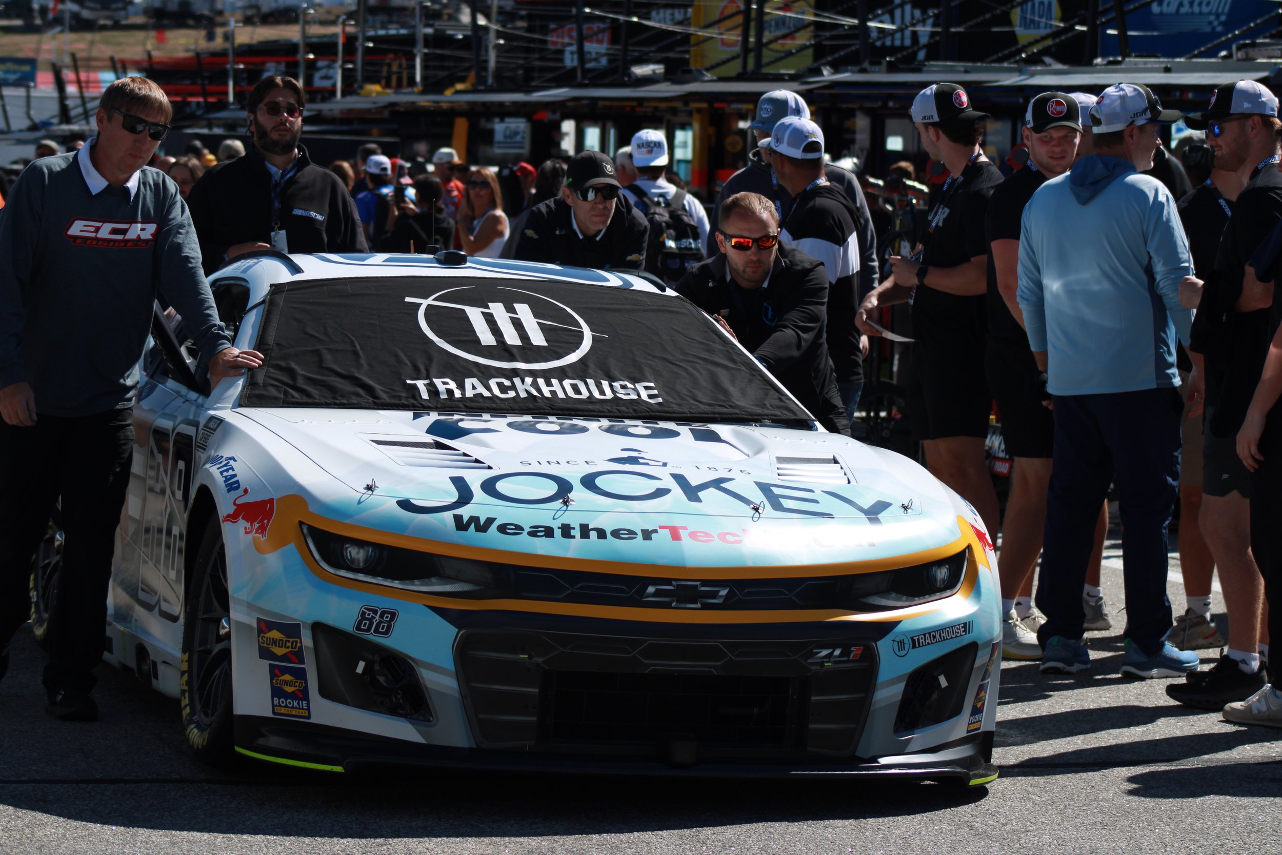 A race car with sponsor logos, surrounded by a team and spectators at a racetrack.