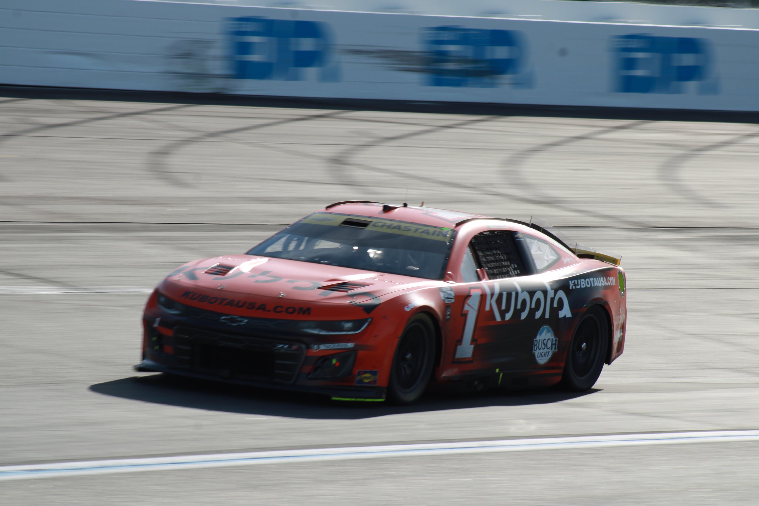 A red race car with black and green accents speeding on a racetrack, sponsored by Kubota with Busch Light logo, with a blue and white wall in the background.