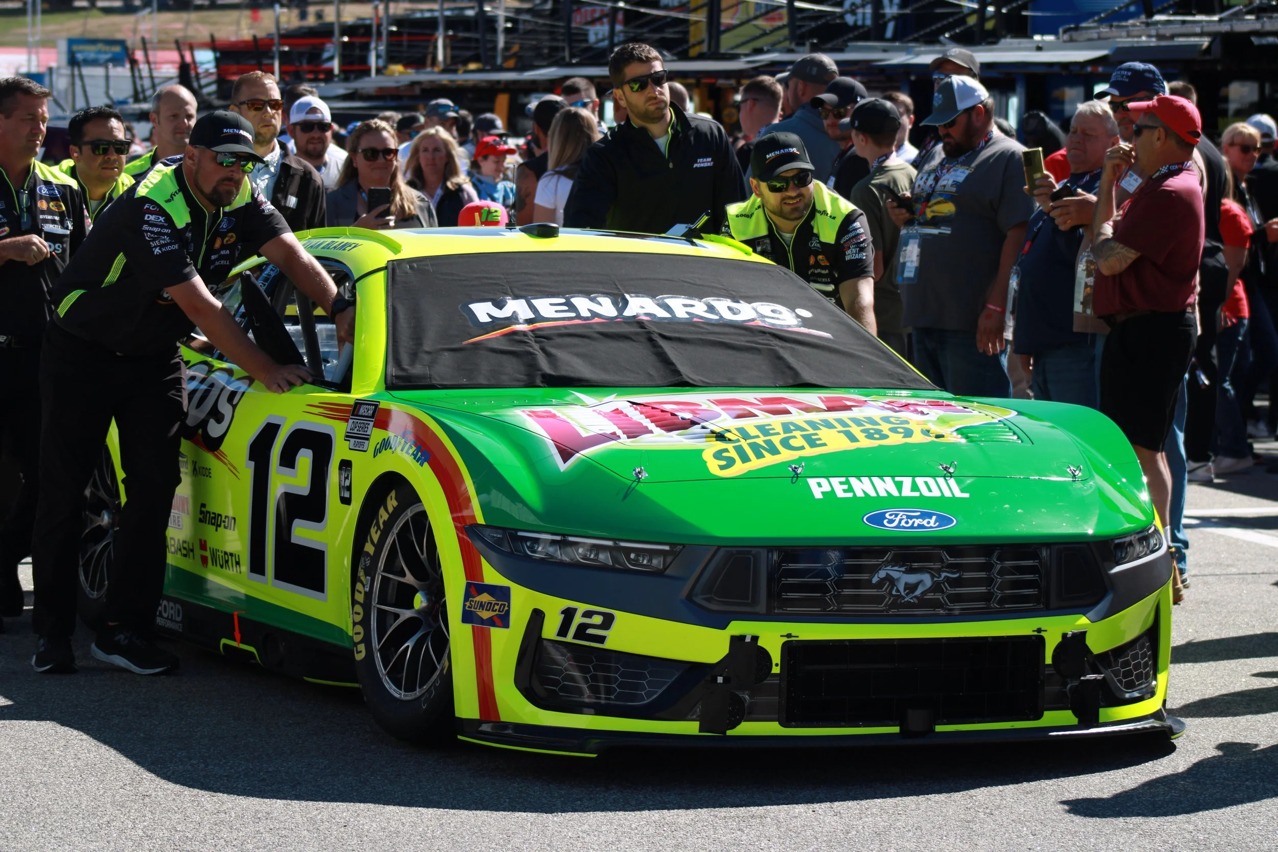 A green and yellow race car with the number 12 and various sponsor logos, surrounded by team members and fans at a racetrack.