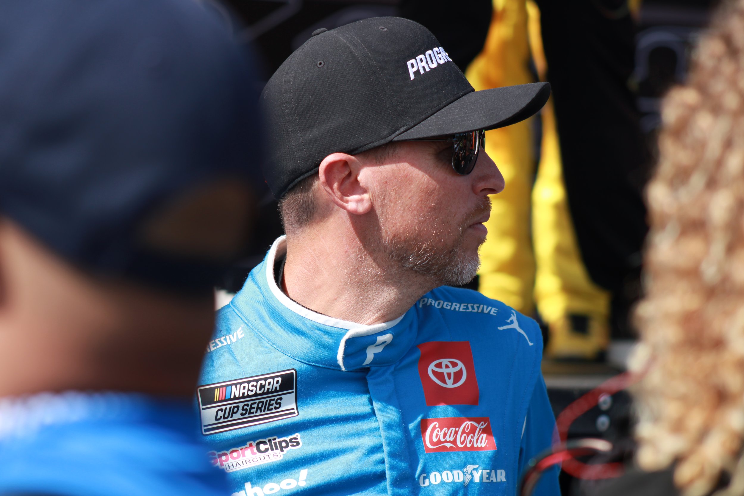 A man wearing sunglasses and a black cap that says 'PROGRESSIVE' in profile view. He is dressed in a blue racing suit with various sponsor logos, including Toyota, Coca-Cola, NASCAR Cup Series, and Goodyear, at a motorsports event.