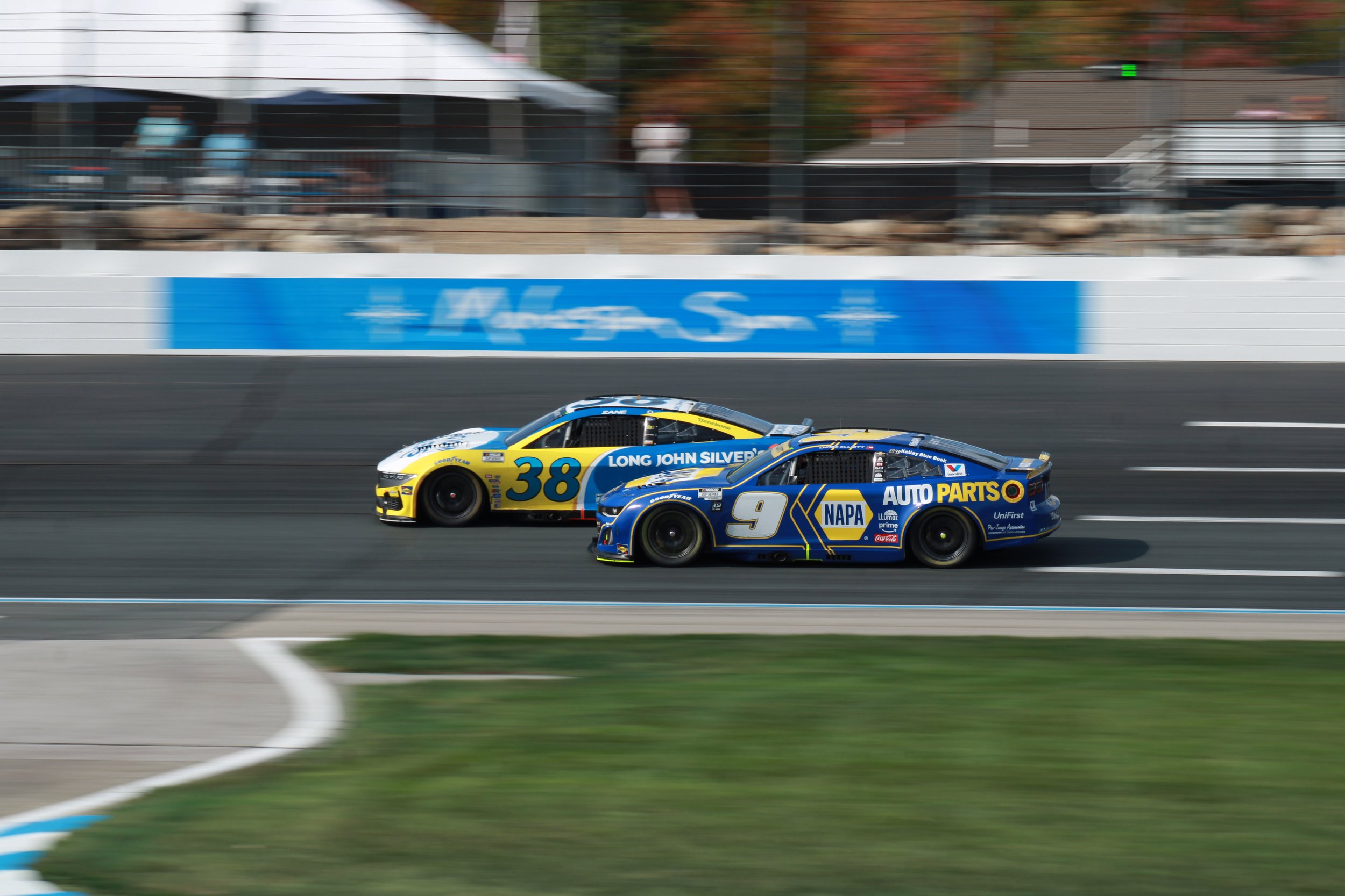 Two race cars speeding on a track, one with the number 38 and the other with the number 9, with sponsor logos such as Napa Auto Parts visible.