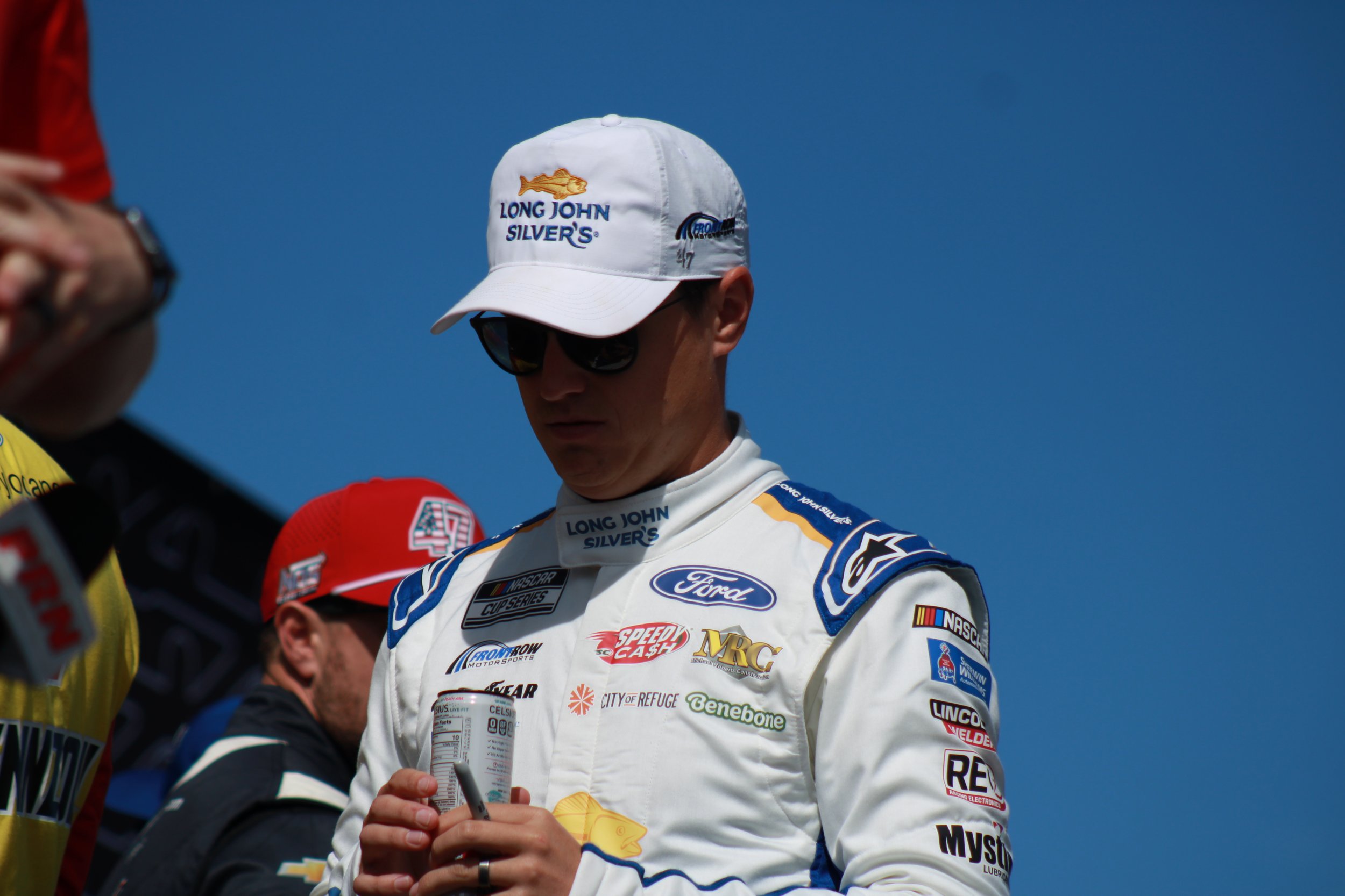 Race car driver in a white racing suit and black sunglasses, wearing a white cap with logos, holding a soda can, standing among others against a blue sky.