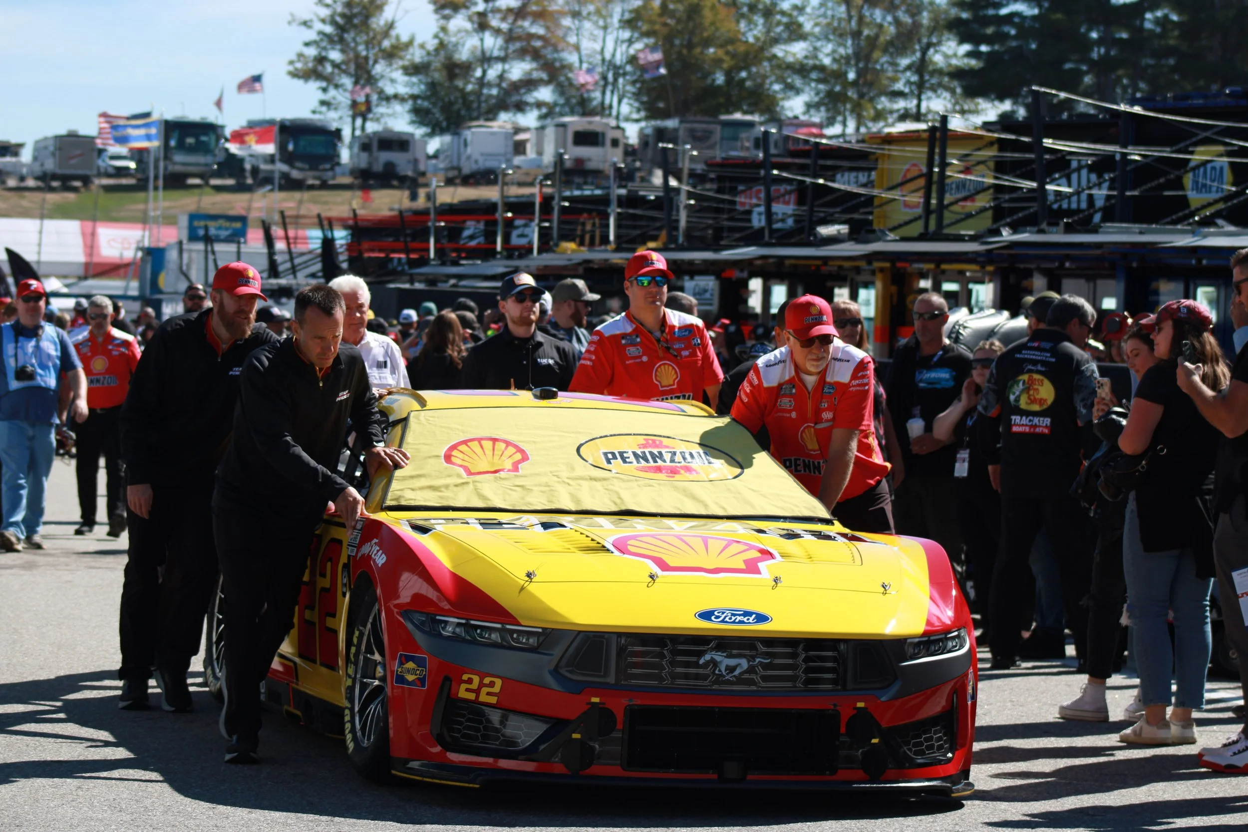 A race car with a yellow and red sponsor logo, the number 22, and a Ford badge is being pushed by a team of mechanics and crew members surrounded by fans and staff at a racing event.