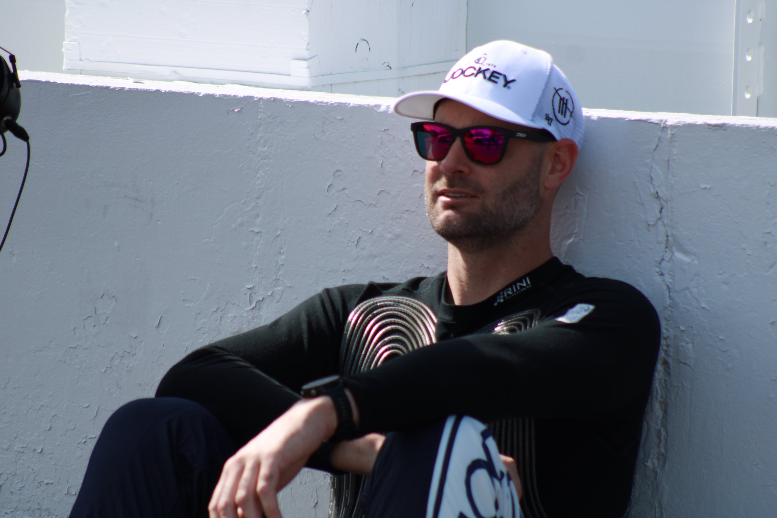 A man sitting against a white wall, wearing a white baseball cap with purple and black text, black sunglasses with pink reflections, a black long-sleeve shirt, and black pants, with his arms crossed.