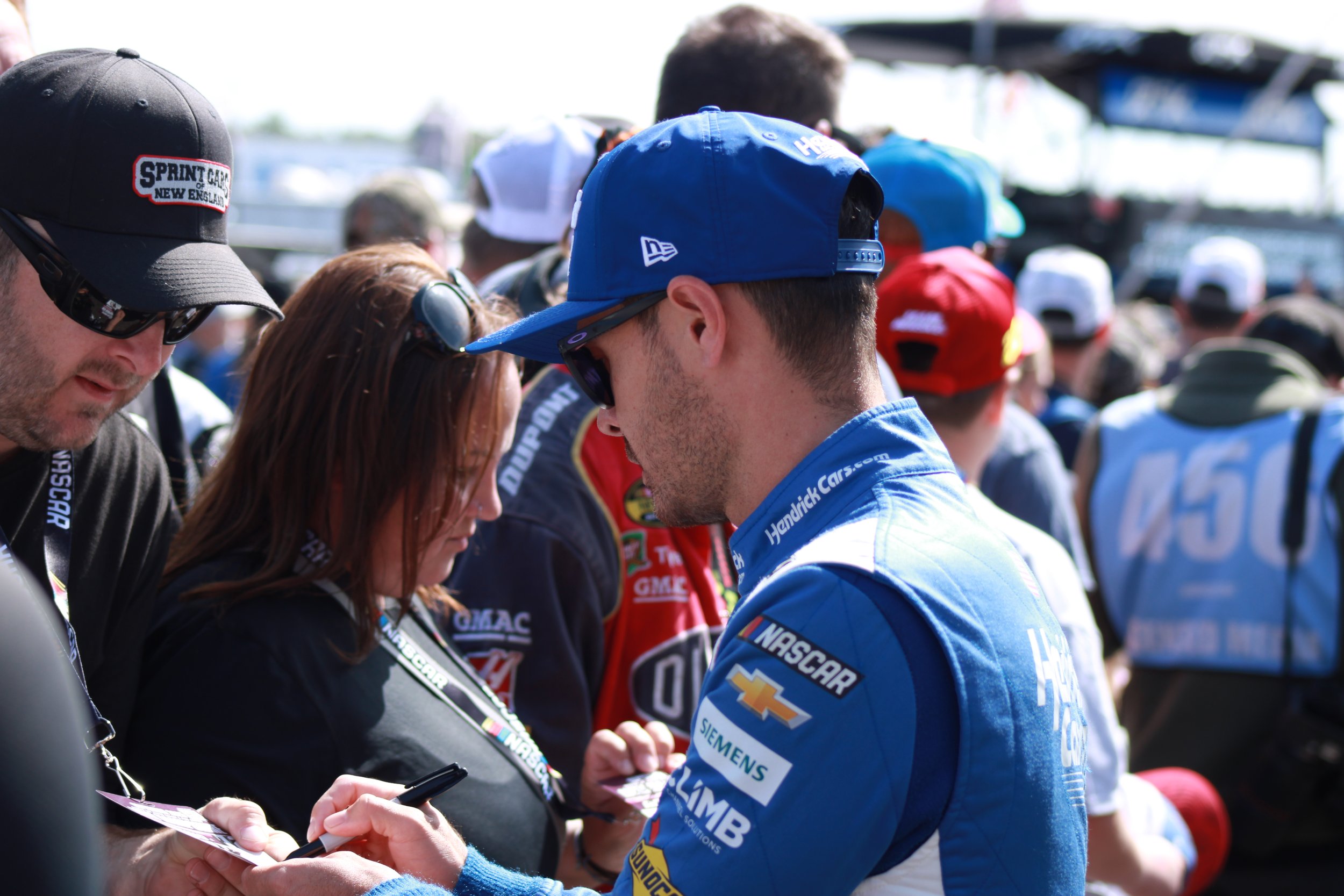 Race car driver in blue HendrickCars.com racing suit signing autographs at a racing event, surrounded by fans and other racers.