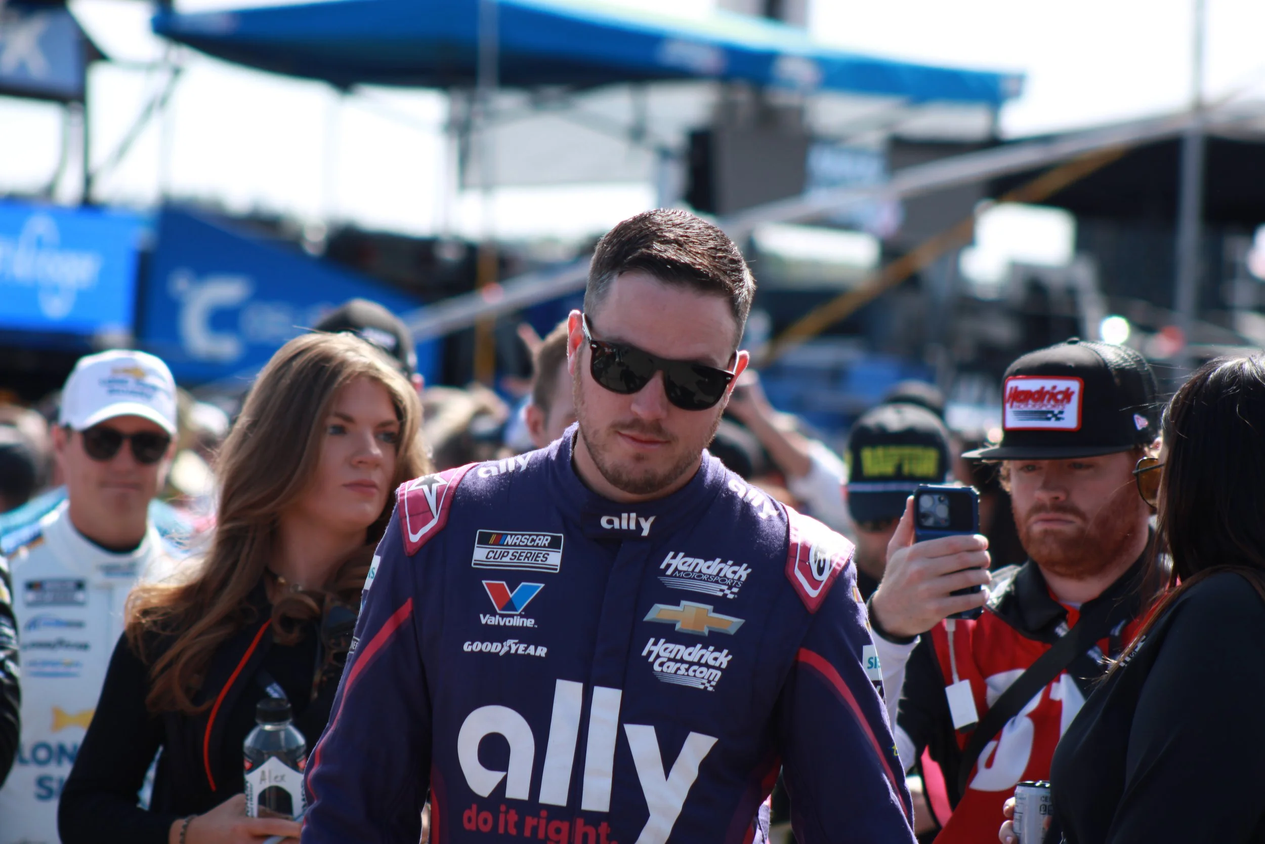 A male race car driver with sunglasses and a racing suit with sponsor logos, surrounded by fans and team members at a racetrack.