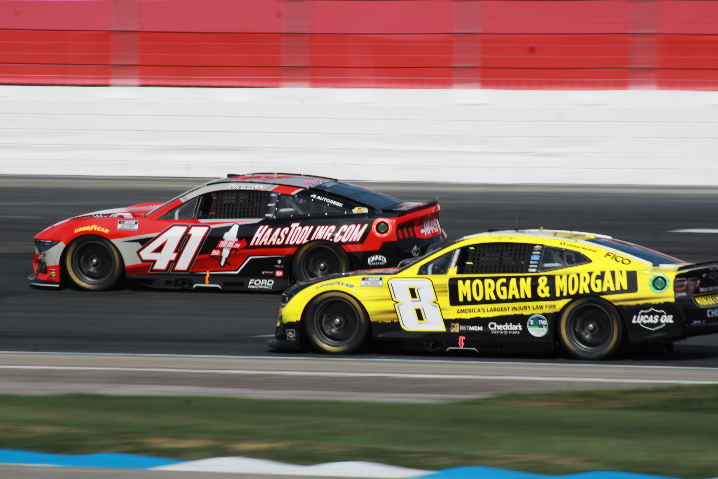 Two race cars are driving on a race track, one red with the number 41 and the other yellow with the number 8, with a red and white wall in the background.