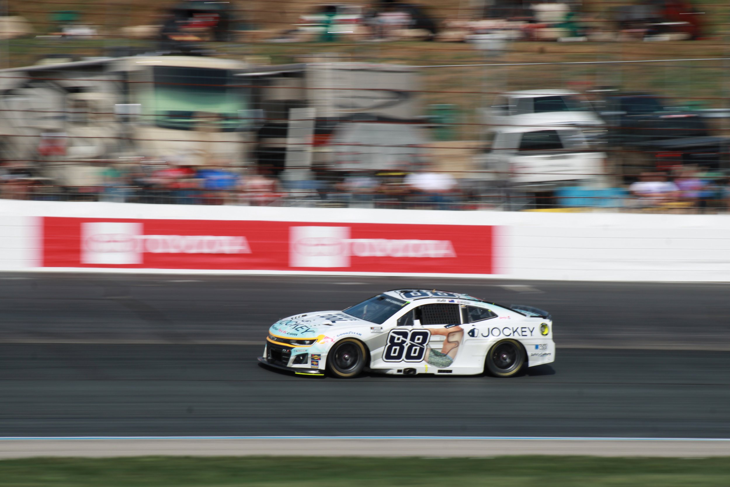 A race car on a racetrack with blurred spectators and vehicles in the background.