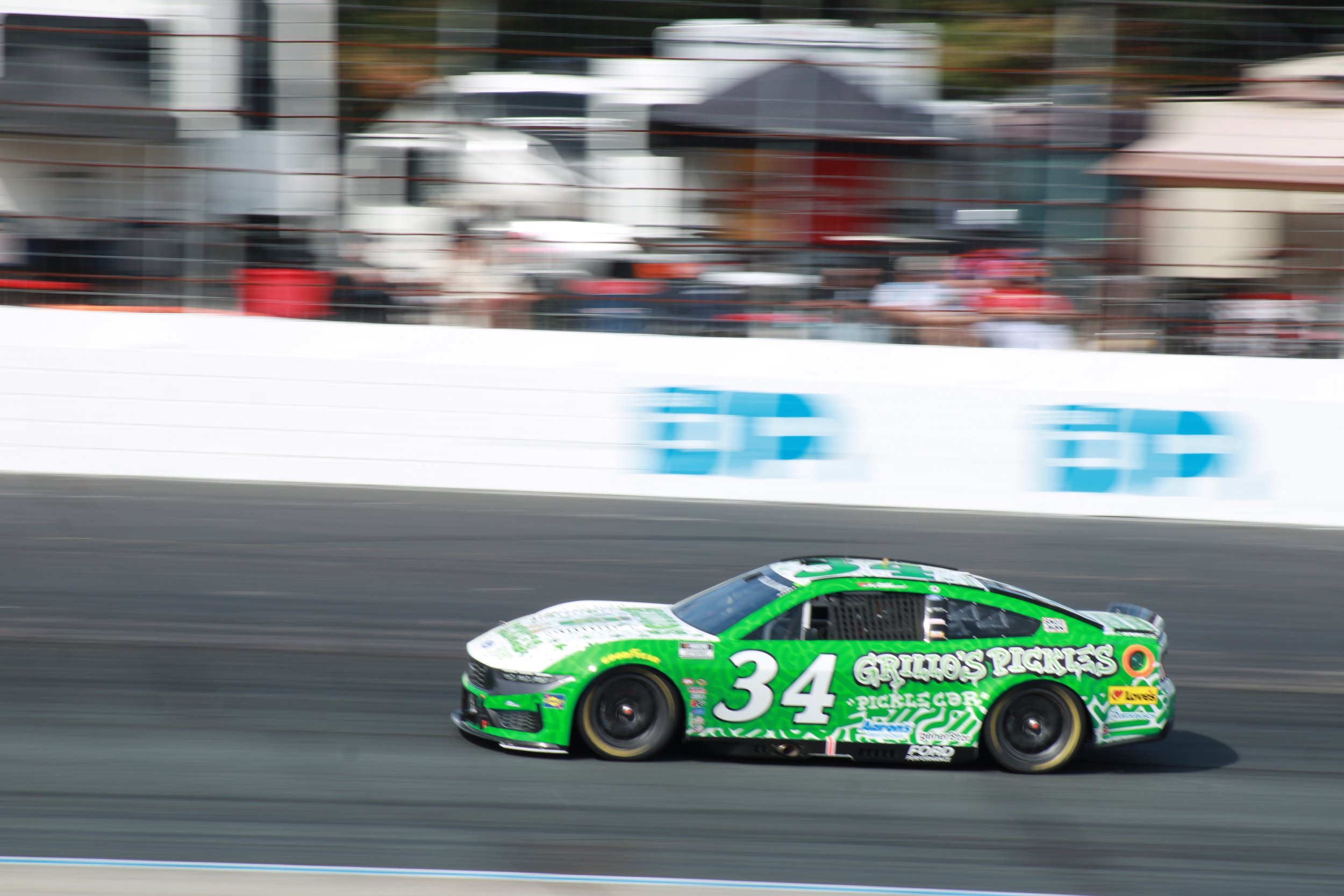 A green race car with the number 34 speeds along a racetrack, with blurred spectators and advertisements in the background.