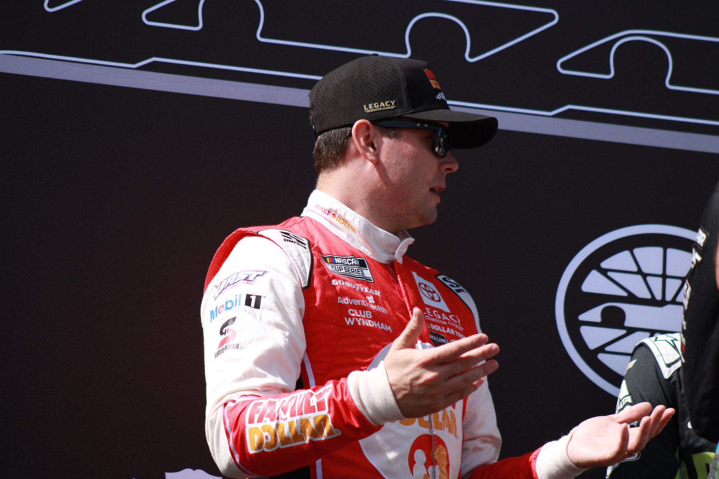 A race car driver in a red and white suit with sponsor logos, wearing a black cap and sunglasses, is gesturing with his hands while standing in front of a black backdrop with a logo.