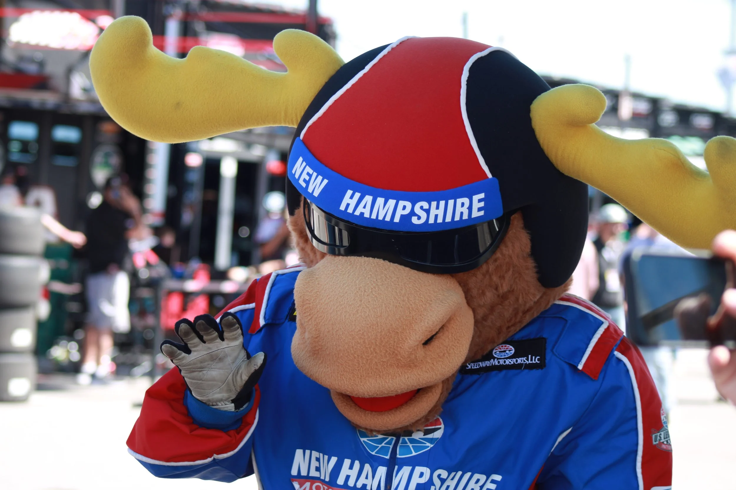 Motocross mascot with a large yellow antler head, wearing sunglasses, a blue racing jacket, and a hat that says "New Hampshire". The mascot is waving and is at a racing event.