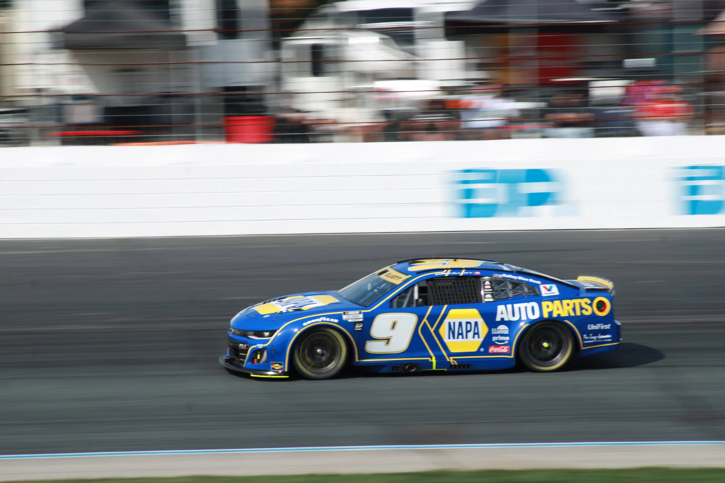 A race car with blue and yellow livery, number 9, moving on a race track with a white barrier and spectators blurred in the background.