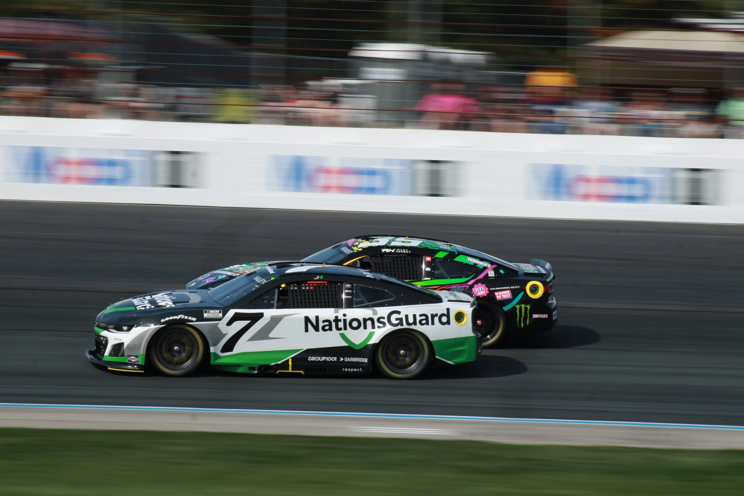 Two NASCAR race cars, one with NationGuard branding and the other with Monster Energy branding, racing on a track with blurred background and spectators watching.