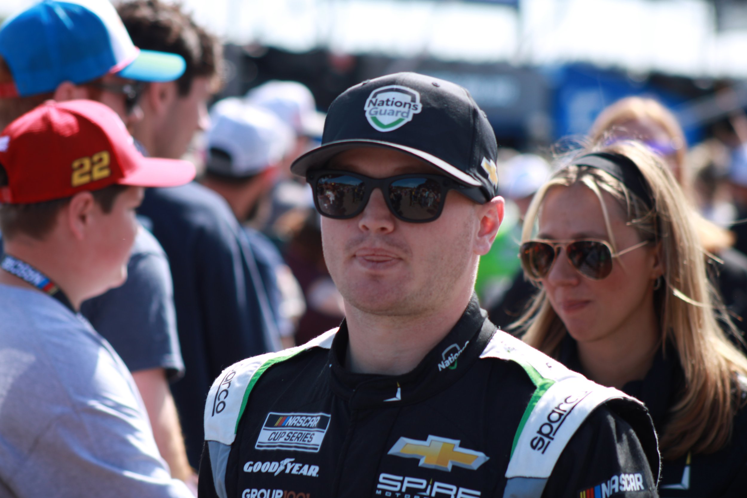 A man wearing a racing suit and sunglasses, with a woman in sunglasses behind him, in a crowded outdoor setting during the daytime.