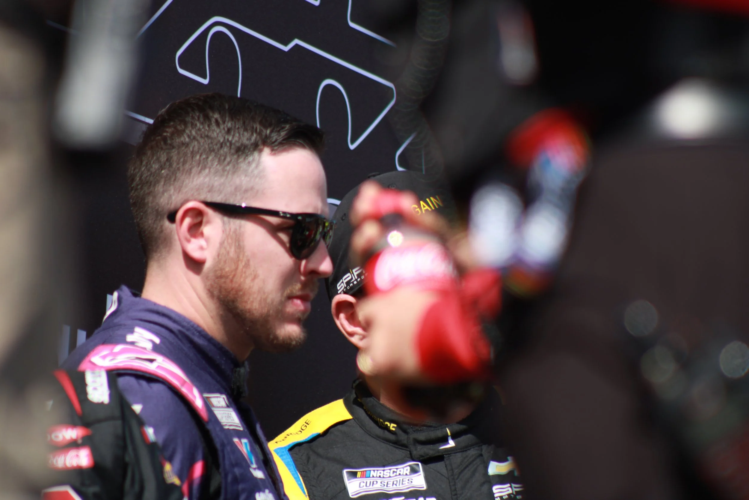 Two NASCAR drivers in racing suits and sunglasses standing close together, with one in the foreground and the other slightly behind, during a race event.
