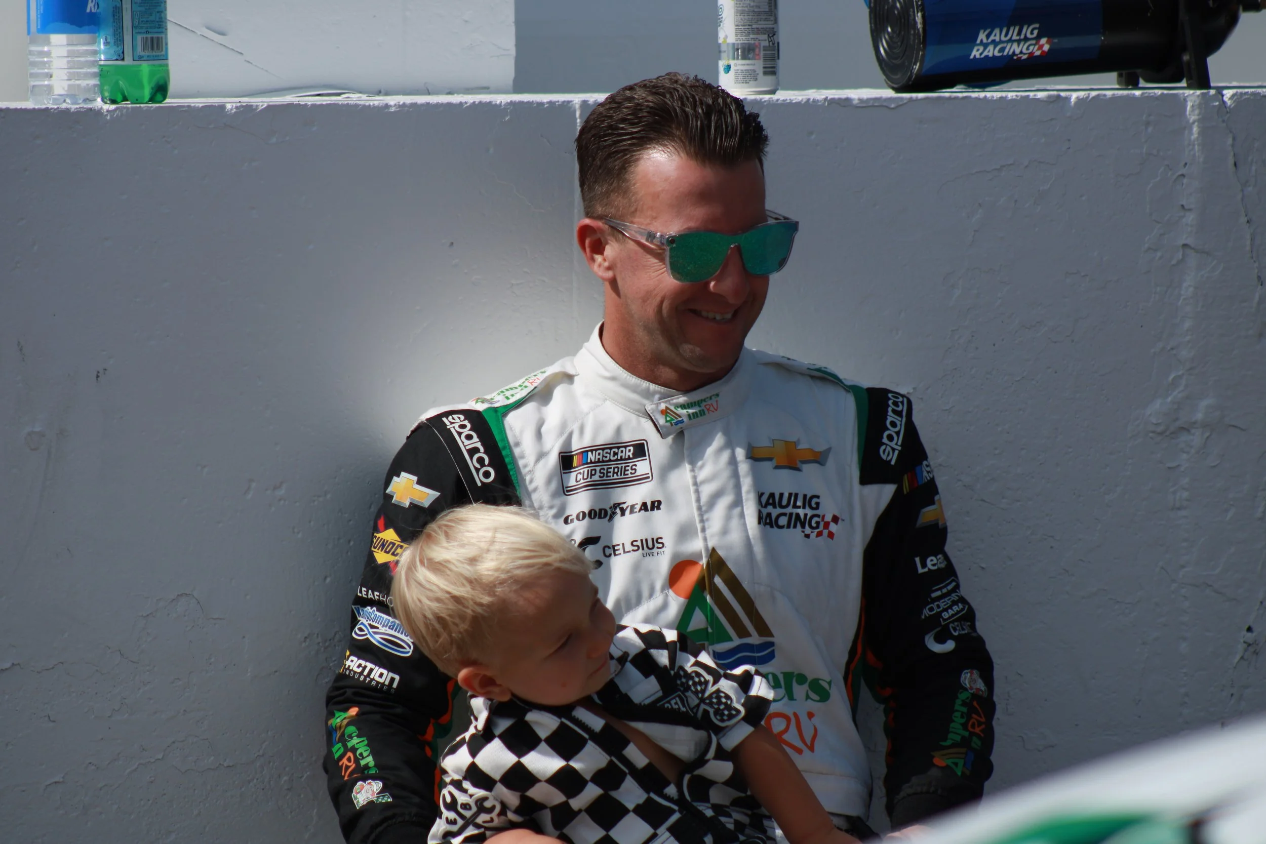 A race car driver in a white racing suit holding a young child, smiling and wearing sunglasses, standing outdoors against a plain white wall.