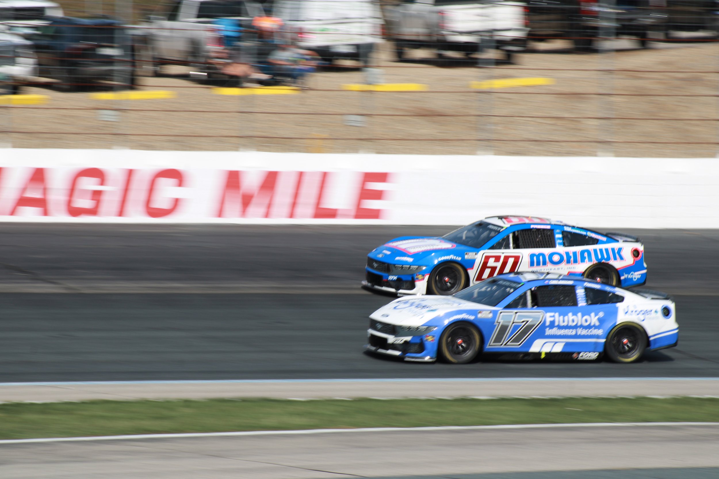 Two race cars, numbered 60 and 17, are on a racetrack during a race. The cars are blue with white and red accents, and they are moving at high speed with a blurred background.