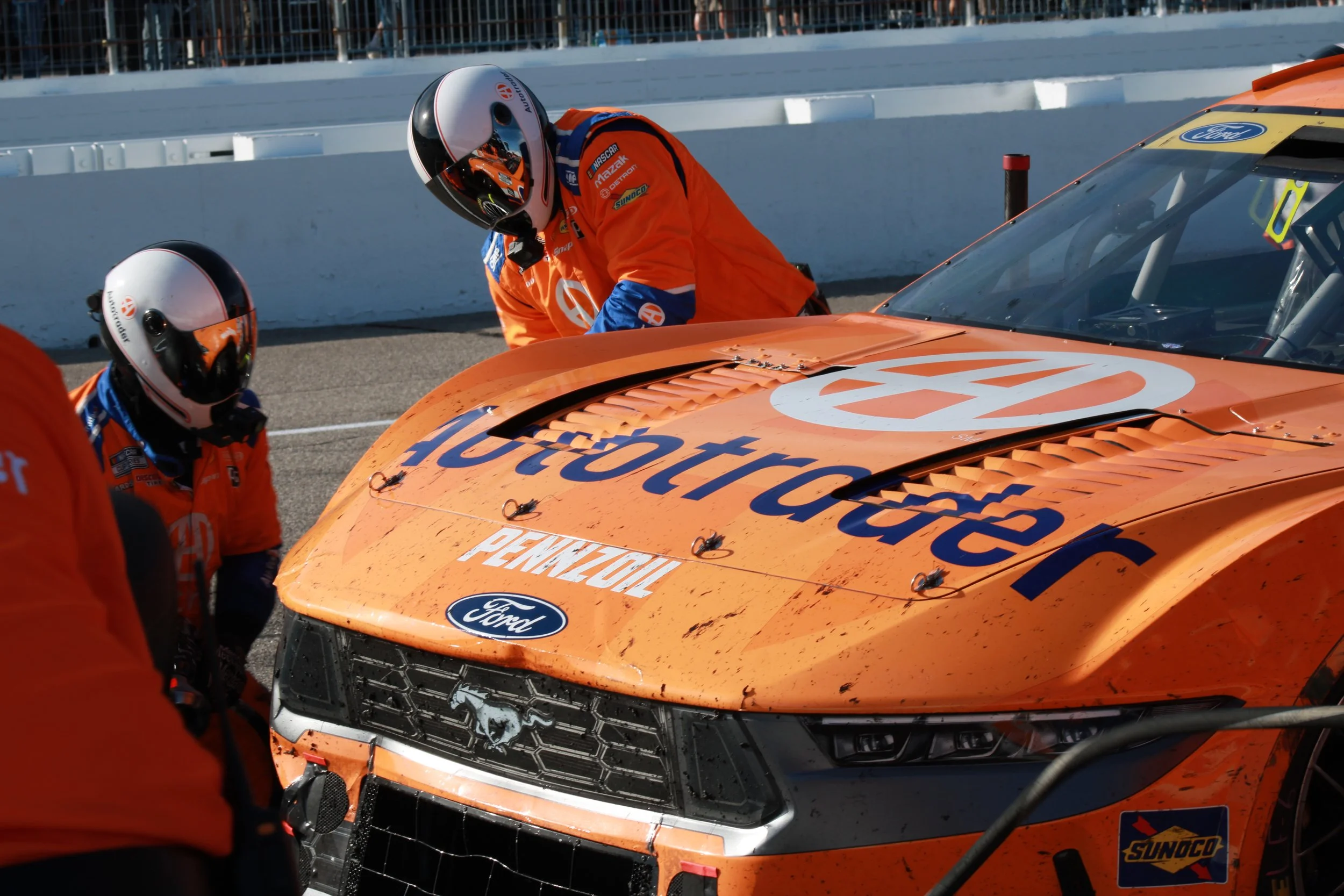 Racecar with sponsor logos and two crew members in orange uniforms and helmets working on it at a racetrack.