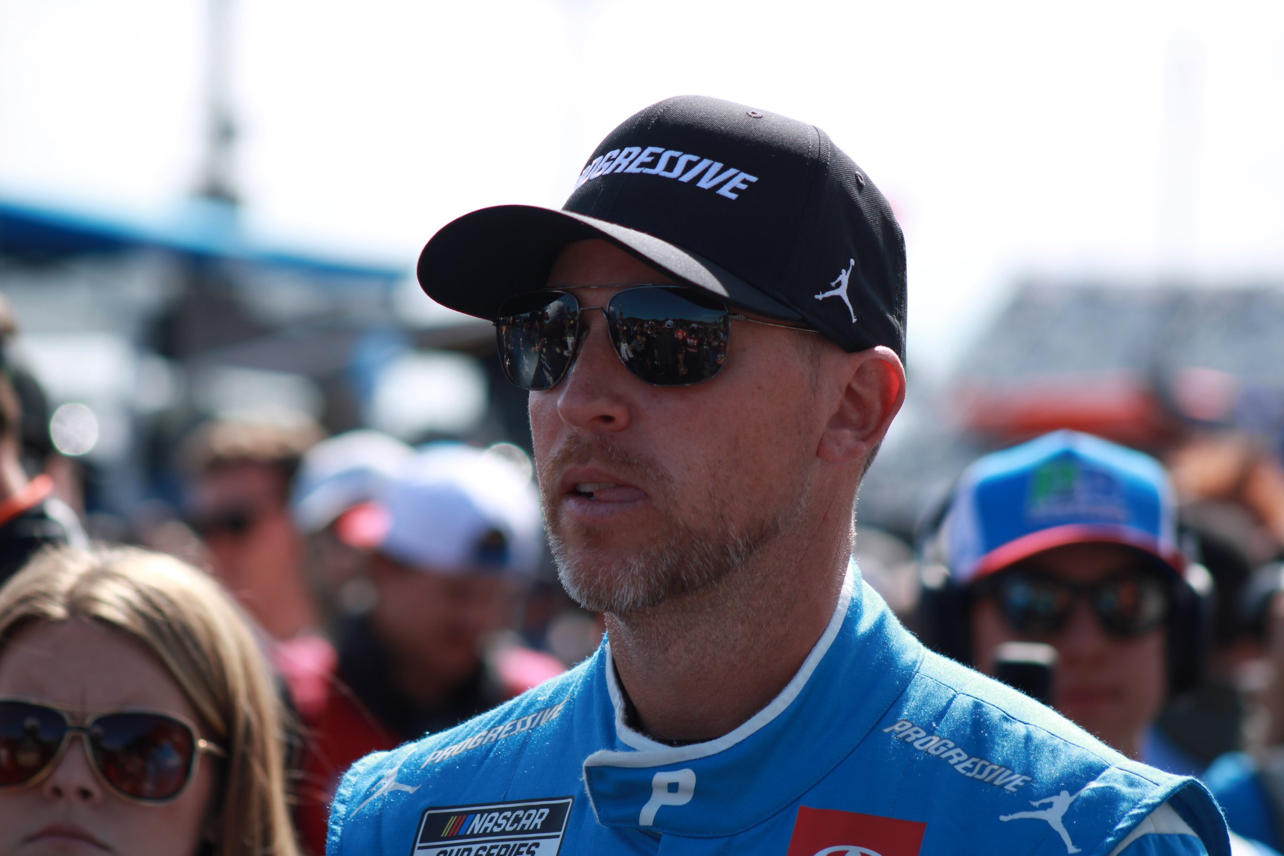 A man wearing sunglasses, a black cap with 'PROGRESSIVE' written on it, and a blue racing suit with sponsors, standing among a crowd at a racing event.