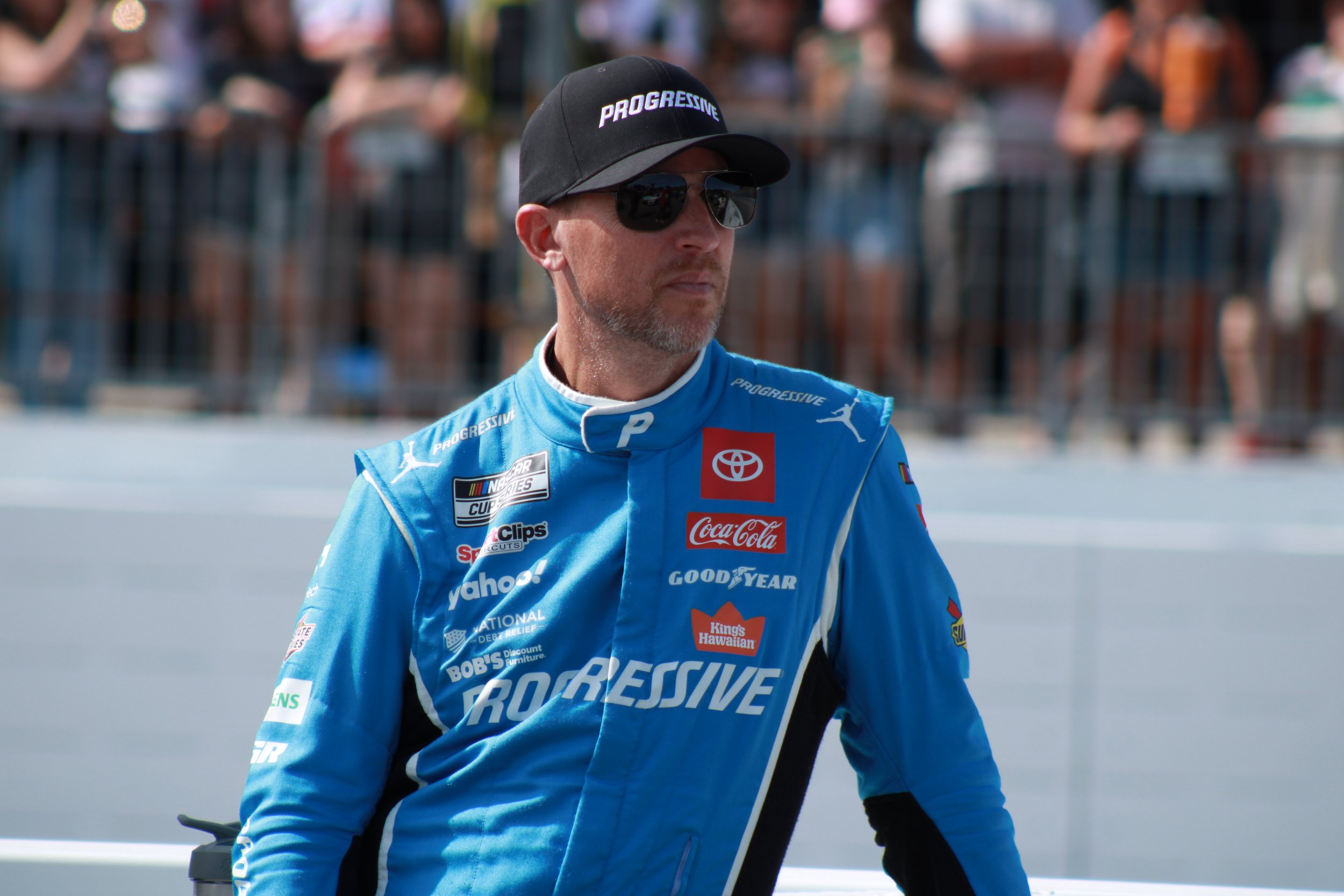 A man wearing a blue racing suit with sponsorship logos, sunglasses, a black baseball cap with 'PROGRESSIVE' written on it, and a white and black collar, standing at a race track with spectators in the background.