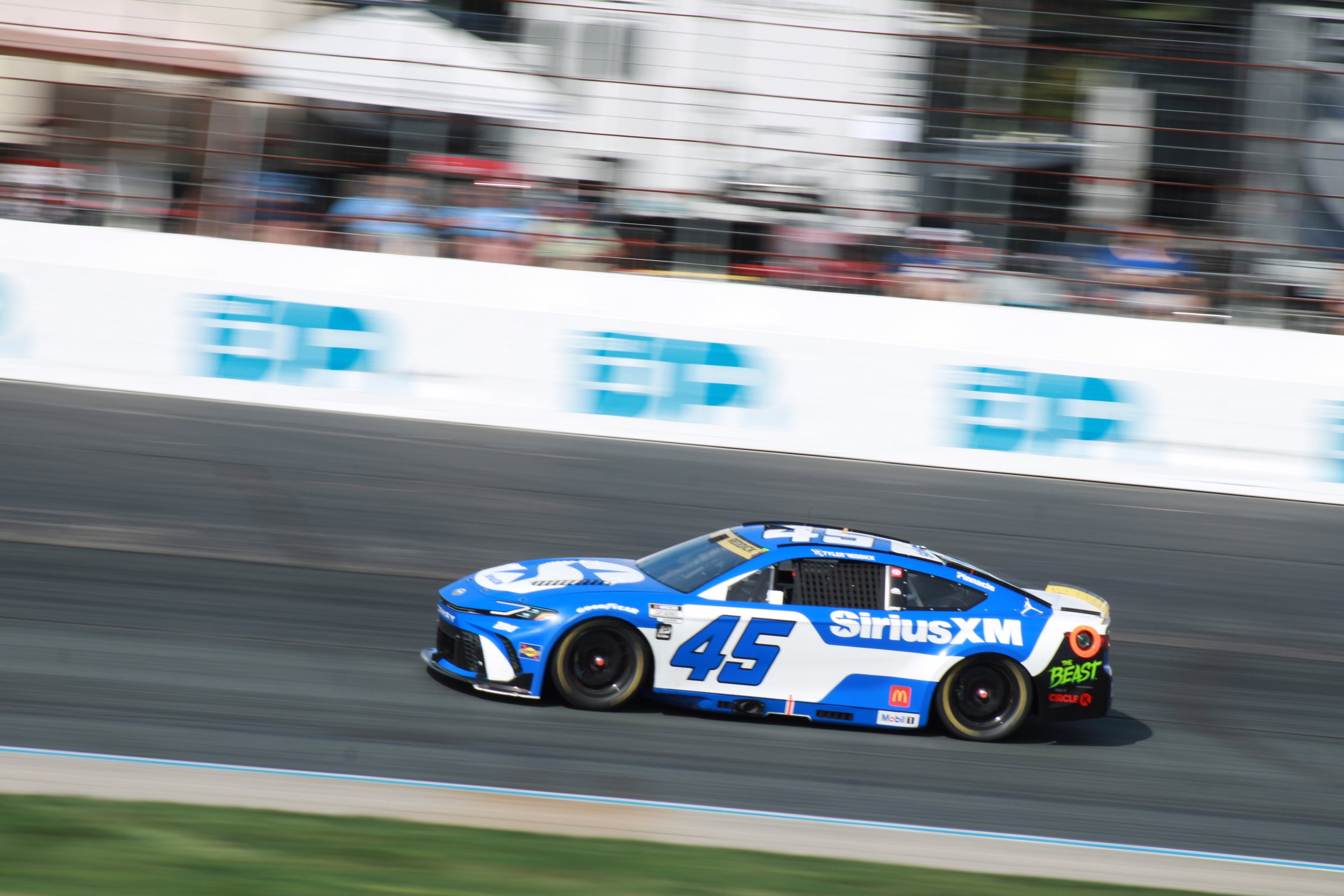 A blue and white stock car with number 45 racing on a track, sponsor logos include SiriusXM and McDonald's, with spectators and barriers in the background.