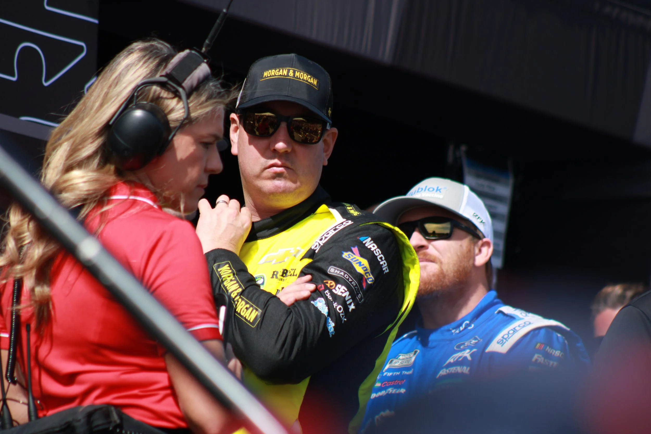 Two race car drivers in racing suits and sunglasses having a serious discussion with a woman wearing a red shirt and headset at a racing event.
