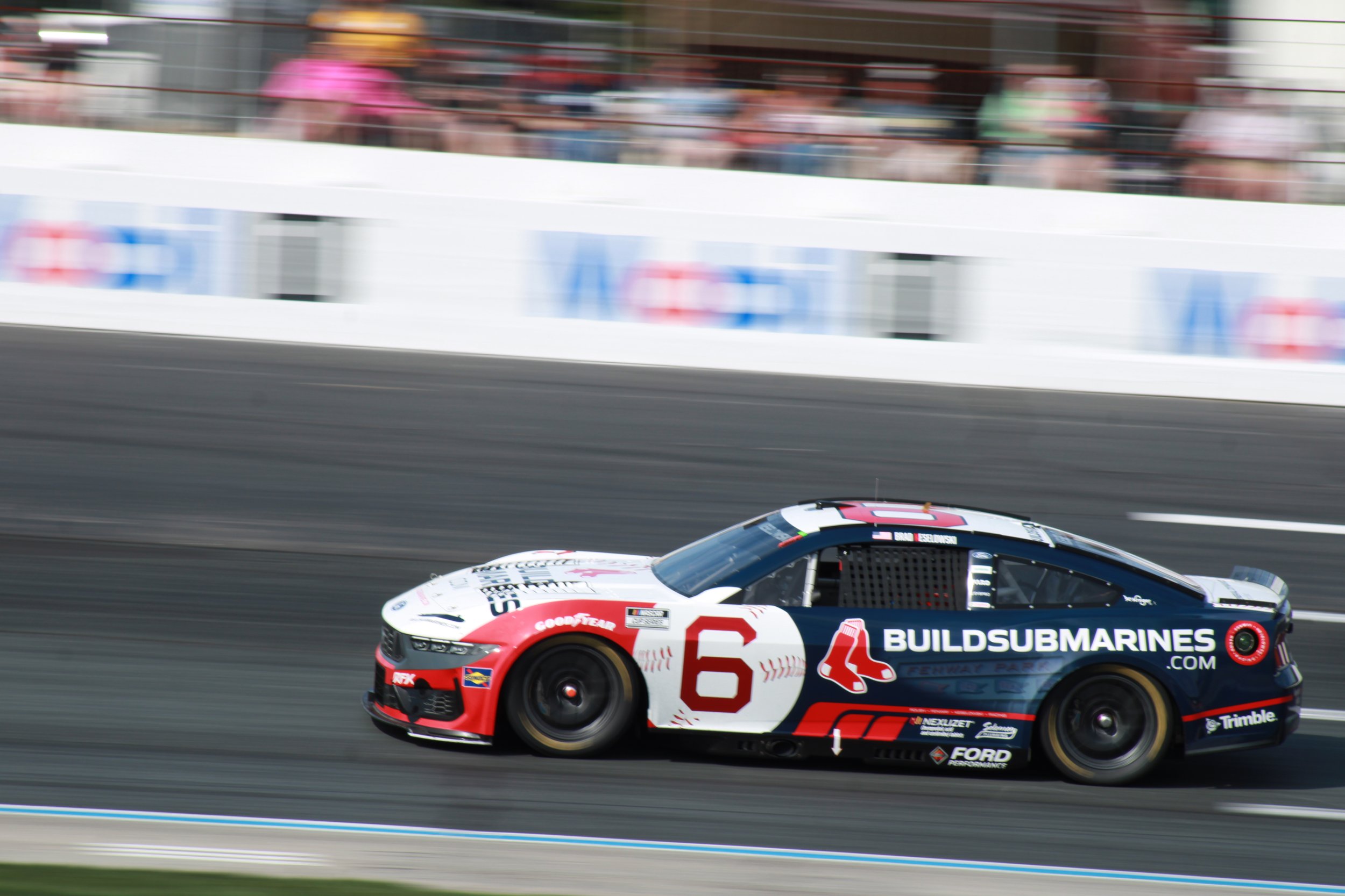 Race car with number 6 speeding on a racetrack, with spectators in the background.