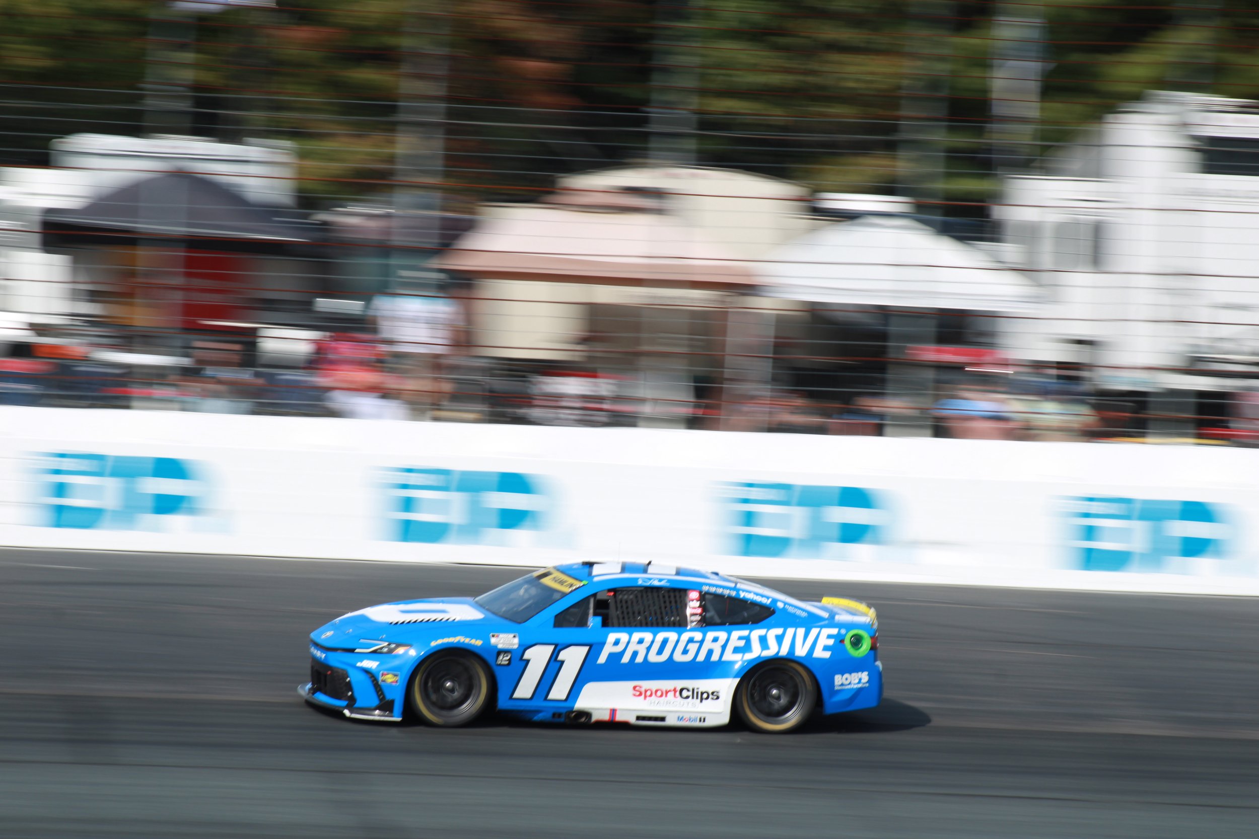 A blue race car with the number 11 and the word PROGRESSIVE on its side speeds along a racetrack, with blurred background and spectators.