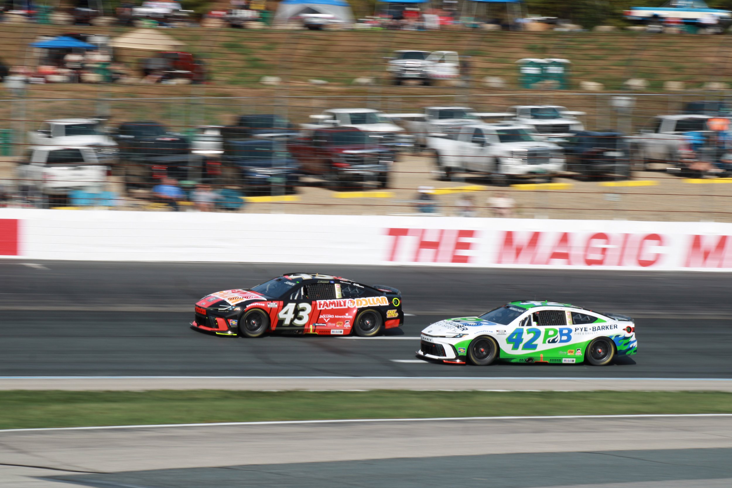 Two race cars, number 43 in black and red and number 42 in white and green, speeding on a racetrack during a race, with a blurred crowd and parked cars in the background.