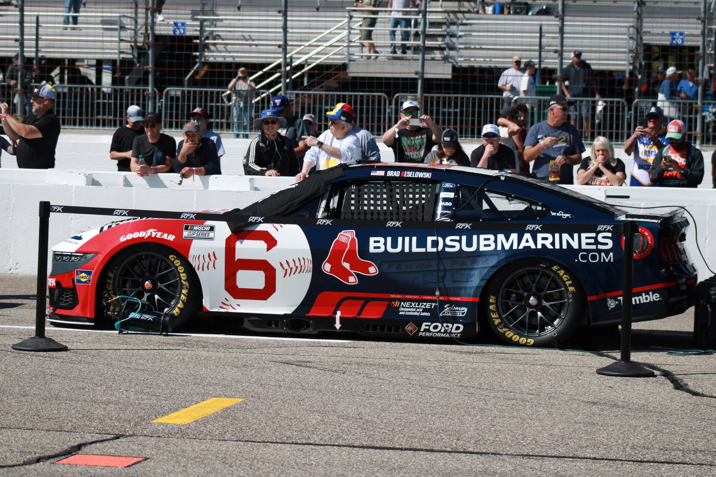 Race car in pit lane with fans watching behind barrier at a racing event.