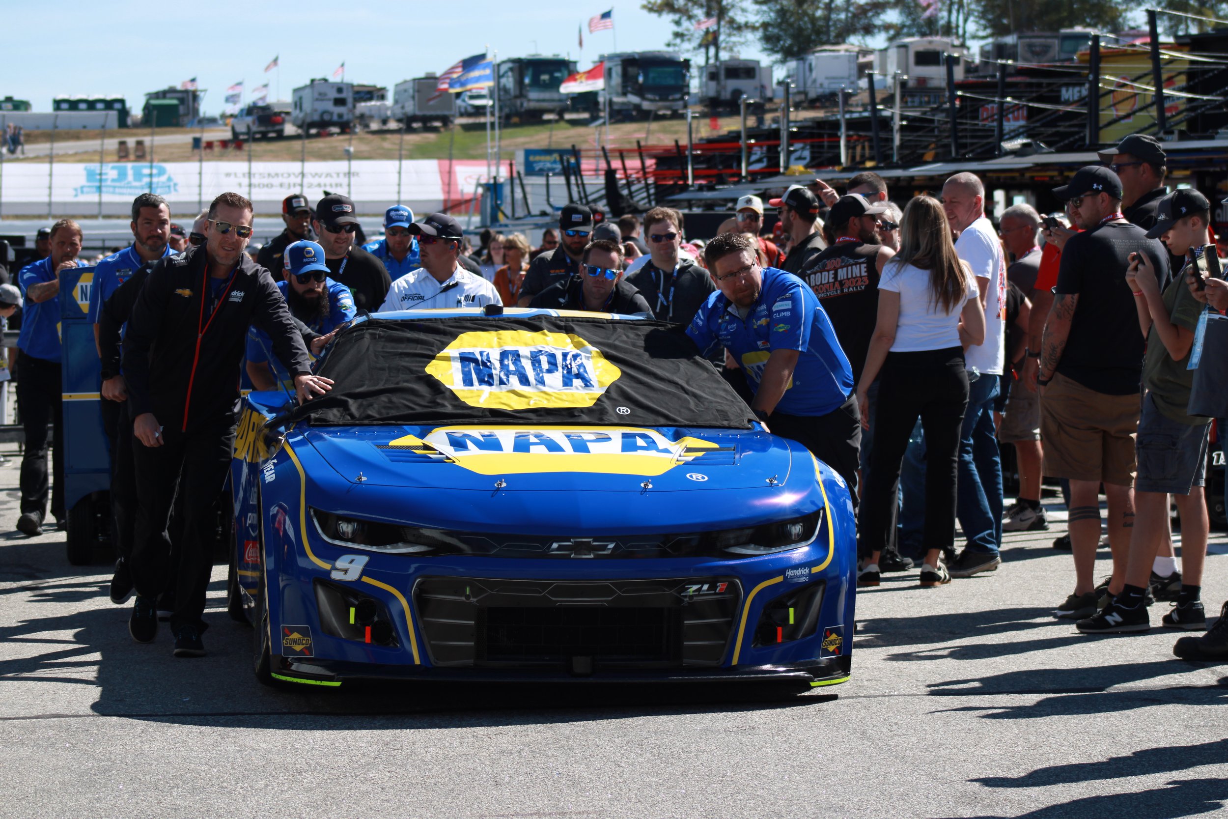 A blue race car with NAPA sponsorship is in the pit area, surrounded by team members and spectators at a racetrack, with flags and trailers in the background.