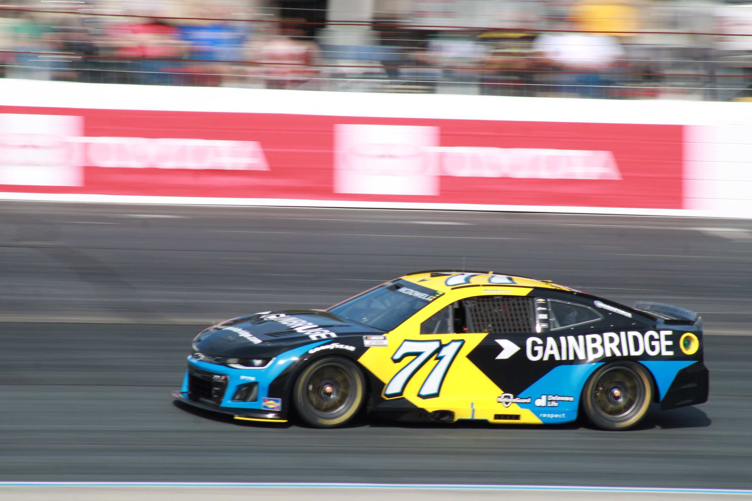 A race car with the number 71 on the side, sponsored by Gainbridge, driving on a race track. The car is black, blue, and yellow with motion blur indicating high speed. Spectators can be seen blurred in the background.