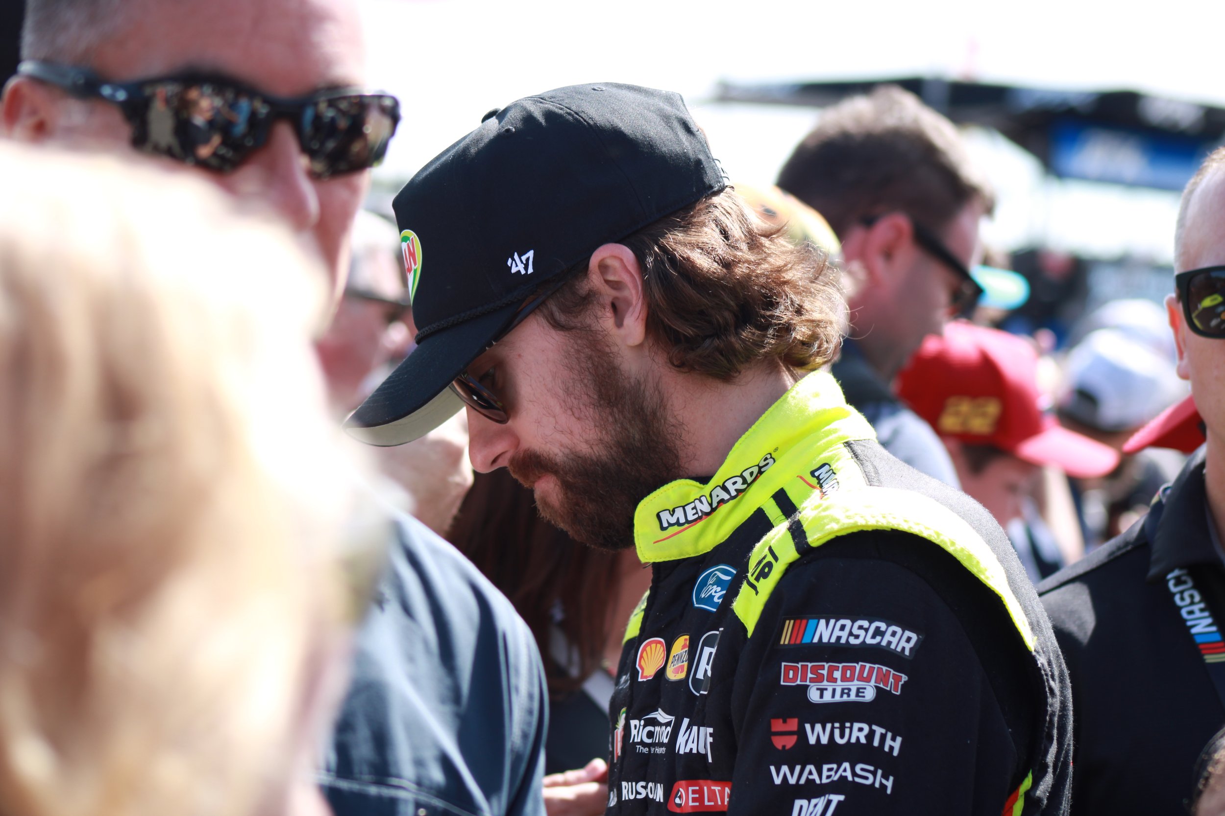 A group of people, including a man in a racing suit and cap, are gathered outdoors. The man in the center has long hair, a beard, and is wearing sunglasses and a bright yellow collar on his racing suit with various sponsor logos. Others around him ar
