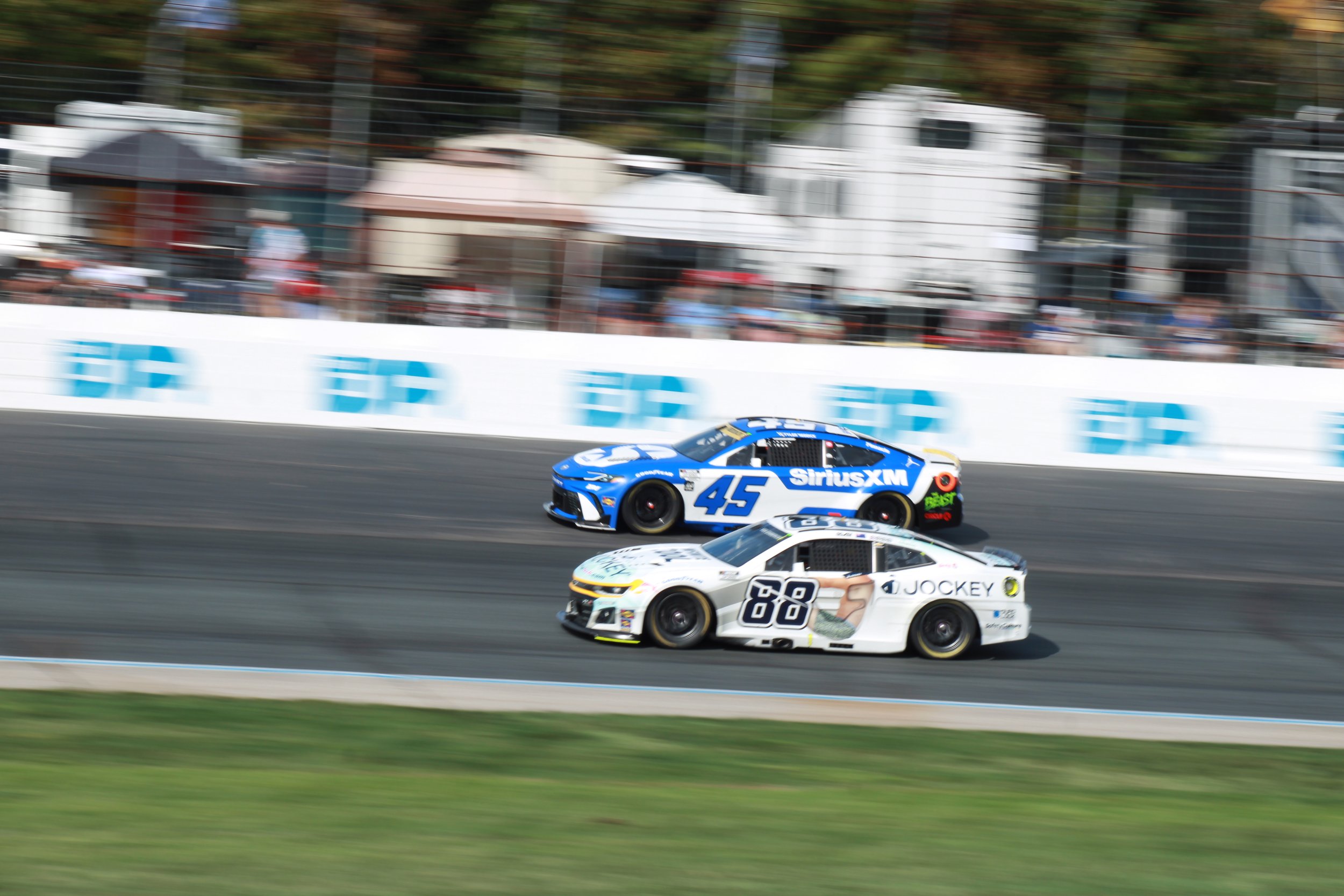 Two race cars, number 45 and number 88, competing on a racetrack with blurred background of spectators and structures.