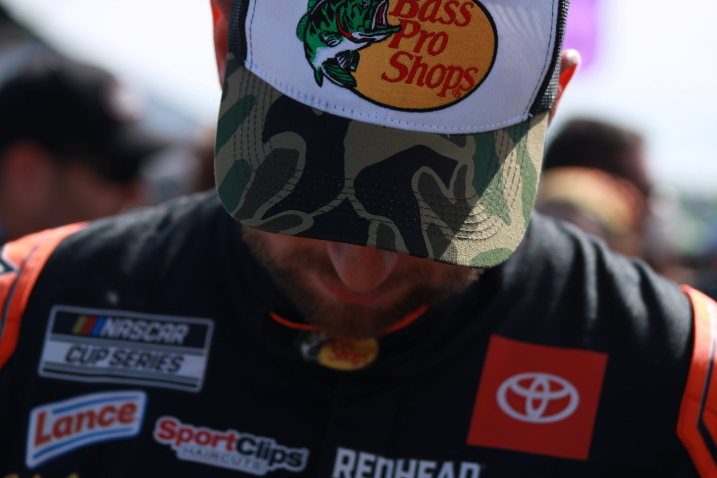 Close-up of a person wearing a racing suit with sponsor logos and a cap with the Bass Pro Shops logo, looking downward.