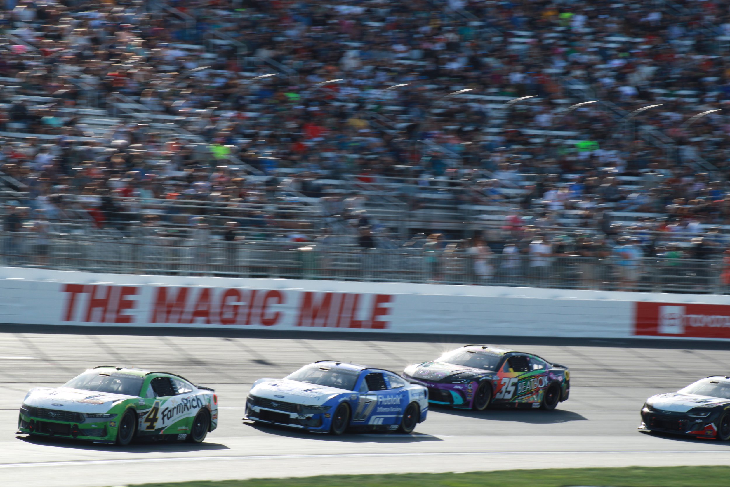 Nascar race cars speeding on the track with crowd in the stands and a sign reading 'The Magic Mile' in the background.