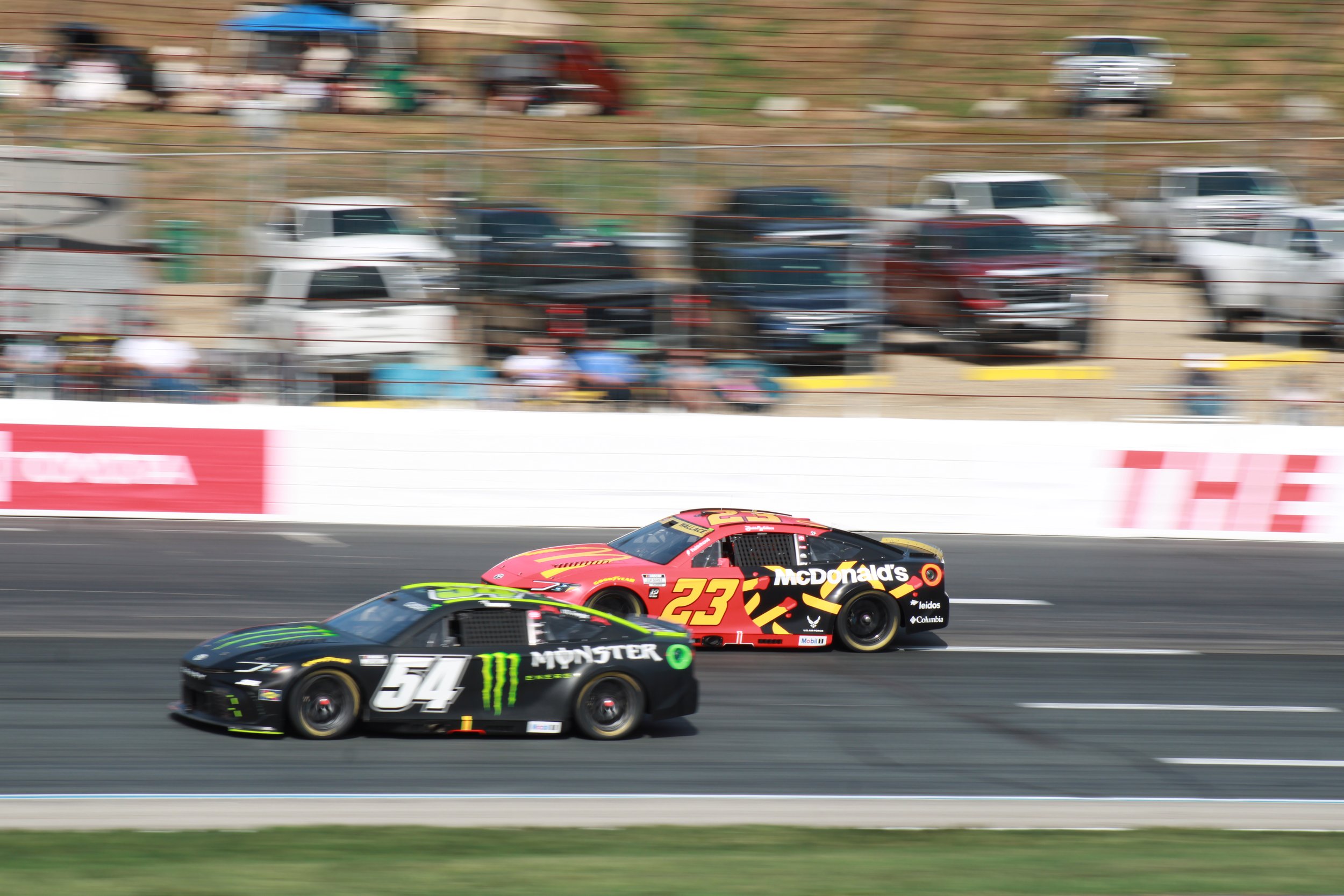 Two race cars speeding on a track, one black with green accents and the number 54, and the other red with yellow accents and the number 23, with a blurred background of spectators and parked cars.