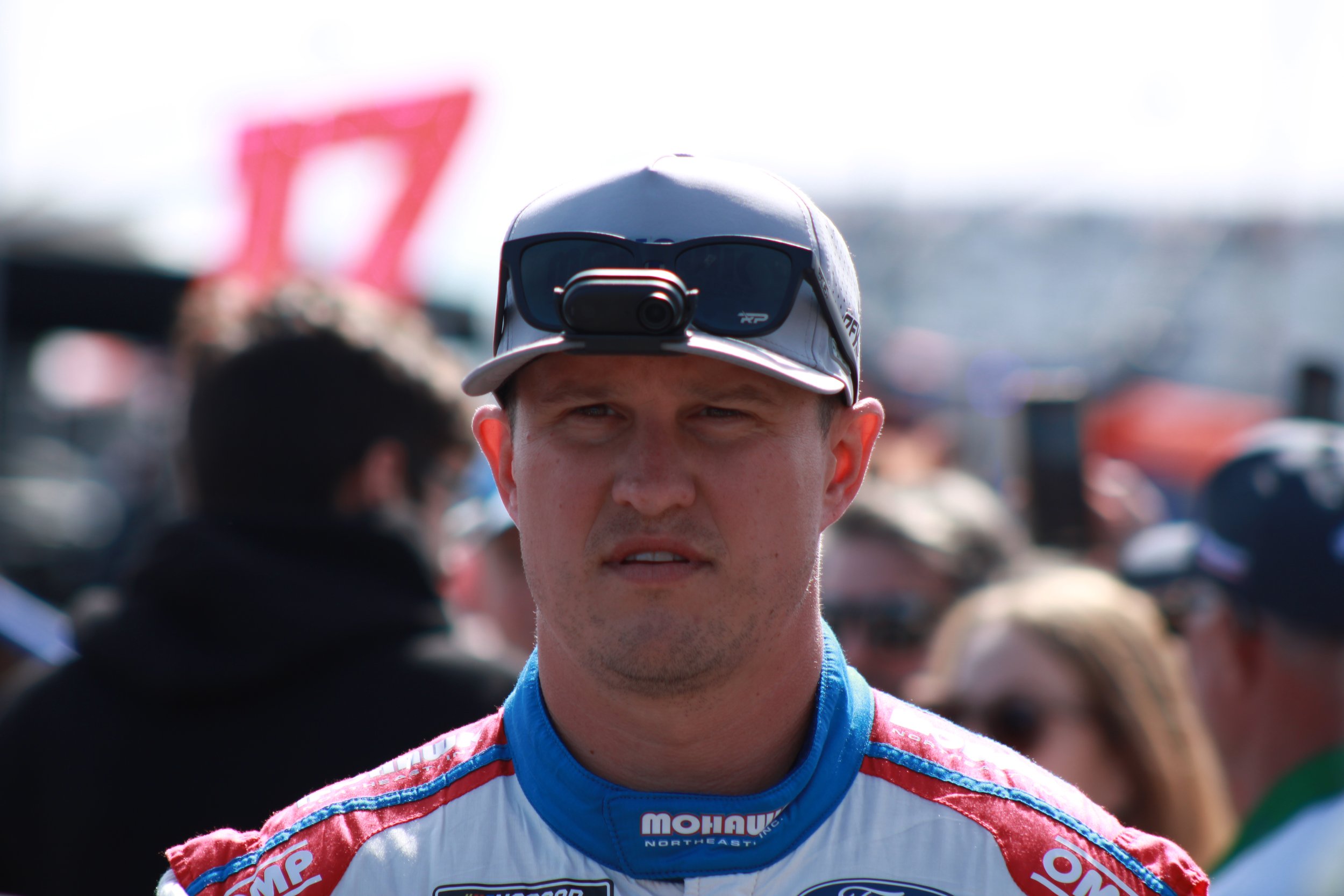 A man in a racing suit wearing a cap and sunglasses with a camera mounted on the cap, at a racing event with a crowd in the background.