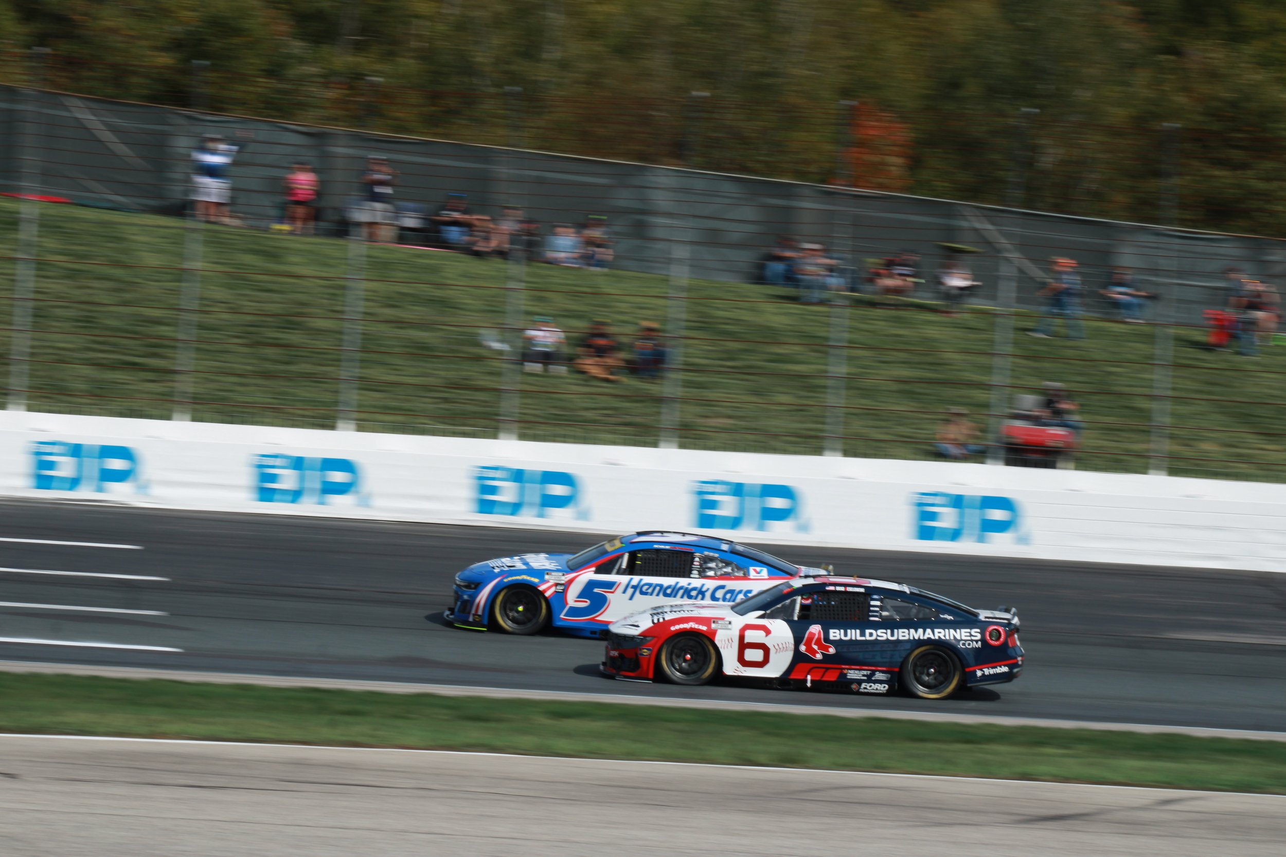 Two race cars, number 5 and number 6, competing on a racetrack with spectators watching from the hillside.