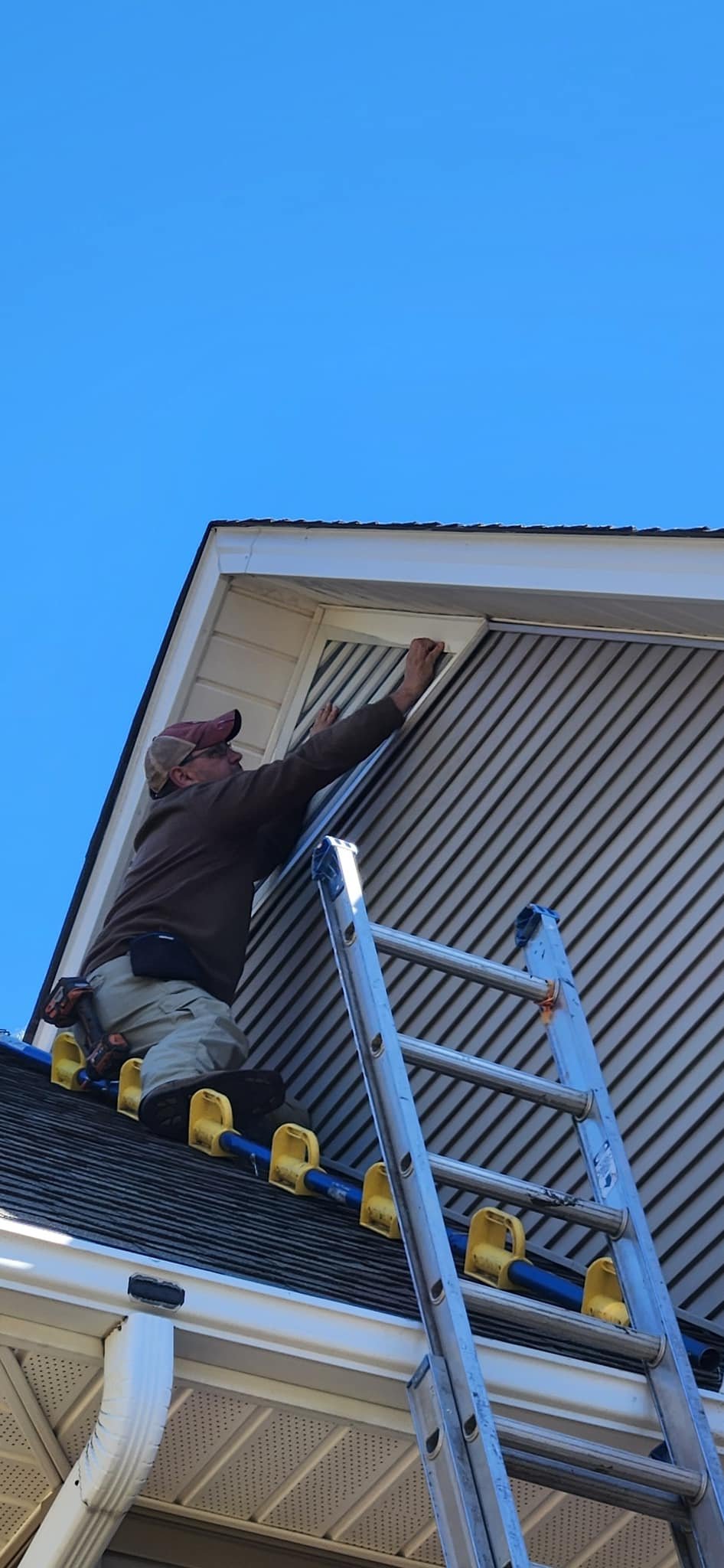 A man installing a gable vent screen on the upper part of a house with a ladder, under a clear blue sky.