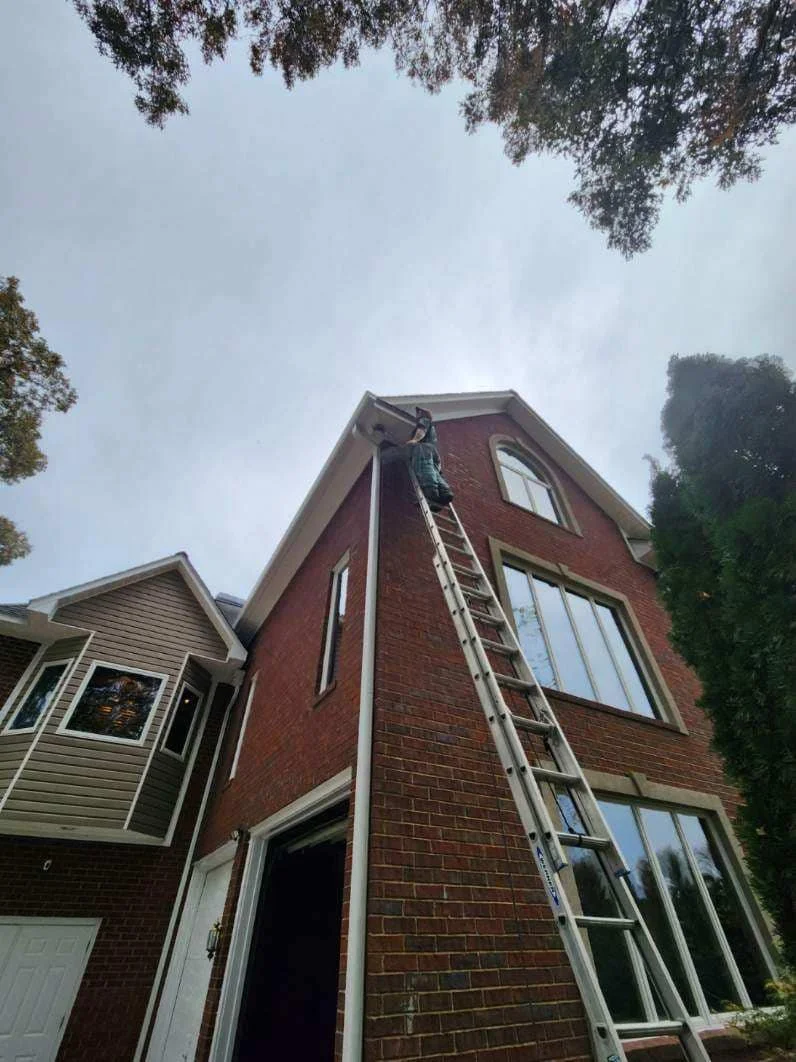 A person climbing a tall ladder leaning against the corner of a red brick house, near the roofline, on a cloudy day.