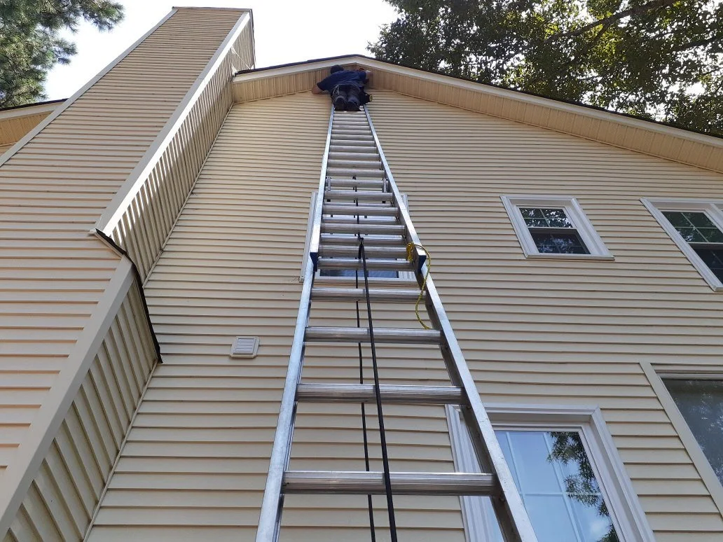 Person climbing a tall extension ladder installing a gable vent screen, near the roofline, with windows and trees visible.