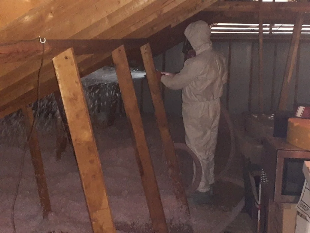 Person in protective suit using a tool on insulation material in an attic.