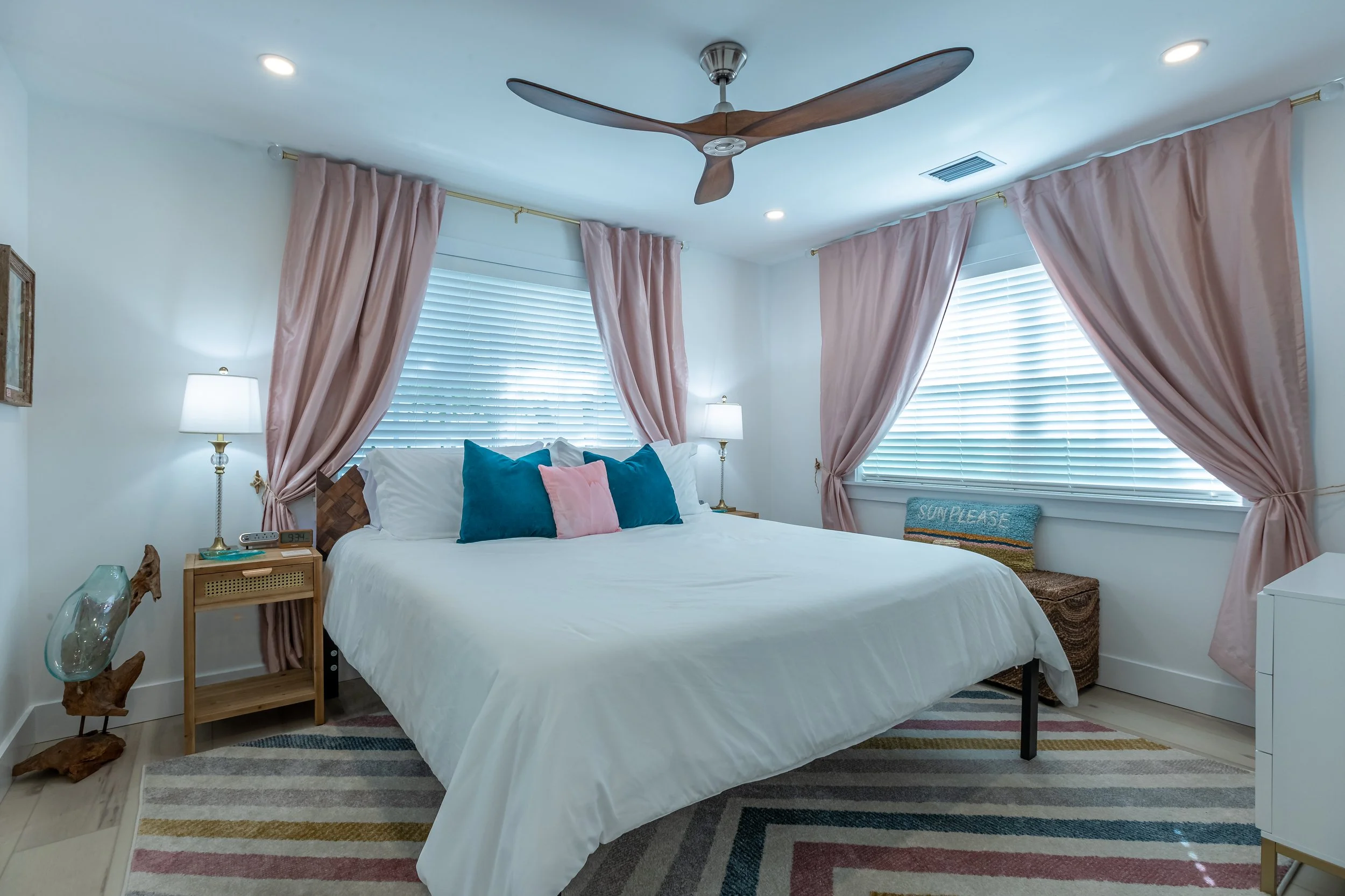 Bright Florida Keys guest bedroom with soft pink curtains, coastal bedding, ceiling fan, and light wood flooring.