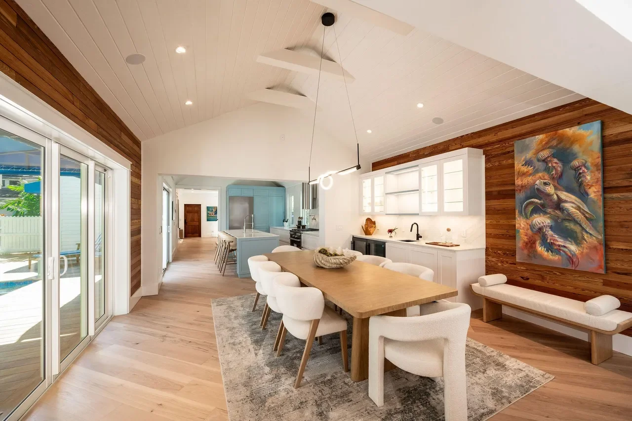 Light-filled Key West coastal dining room with Dade county pine paneling, white shiplap ceiling, wide plank wood floors, modern upholstered chairs, and sliding glass doors opening to a tropical outdoor pool.