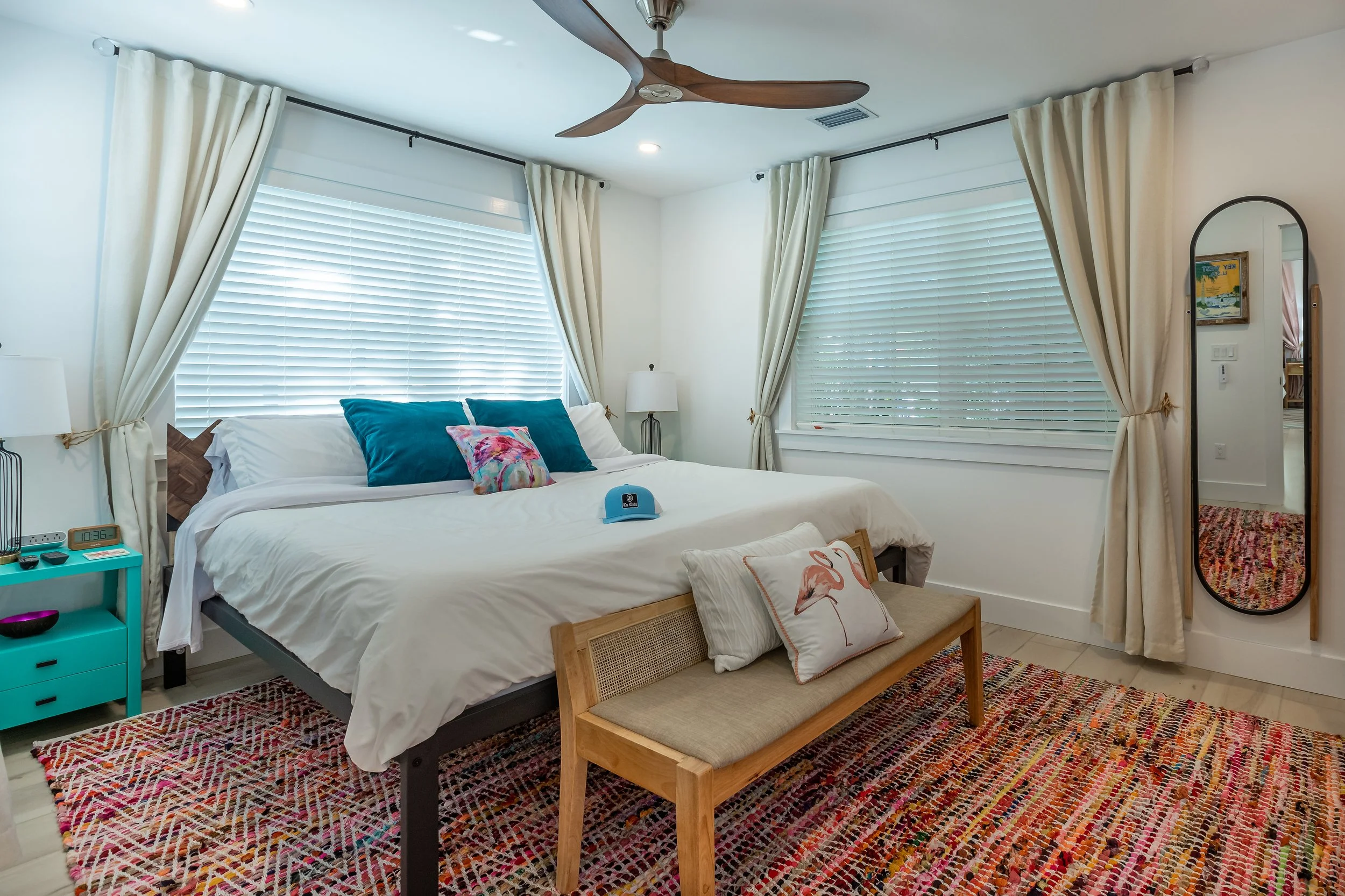 Colorful Key West bedroom featuring layered rugs, white bedding, coastal accent pillows, ceiling fan, and relaxed island style.