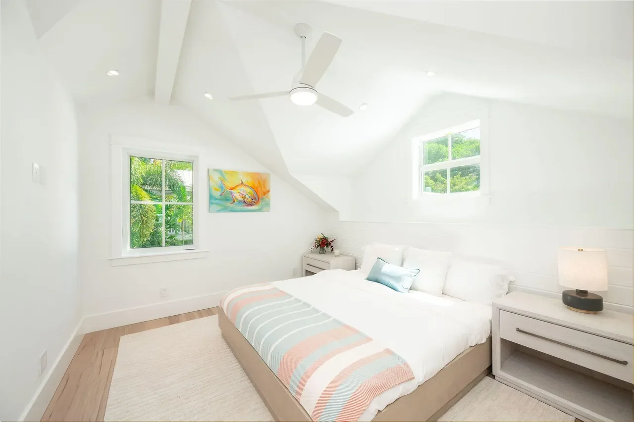 Serene Florida Keys bedroom with vaulted ceiling, coastal-inspired bedding, soft neutral palette, ceiling fan, and windows overlooking palm trees.