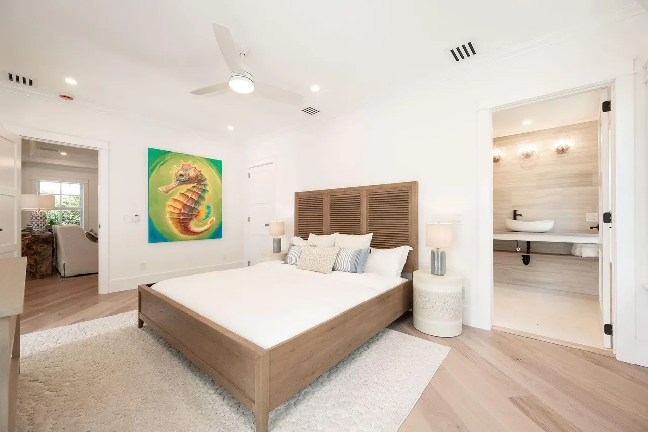 Serene Key West coastal bedroom with a custom wood bed, white bedding, natural wood headboard, coastal artwork, light wood floors, and an open en-suite bathroom designed for relaxed island living.