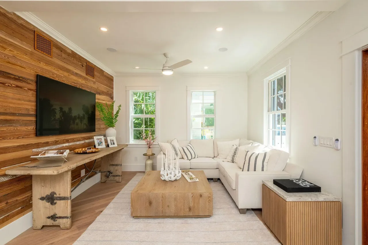 Modern Key West sitting room with custom Dade county pine media wall, white sectional sofa, natural textures, and large windows overlooking tropical landscaping.