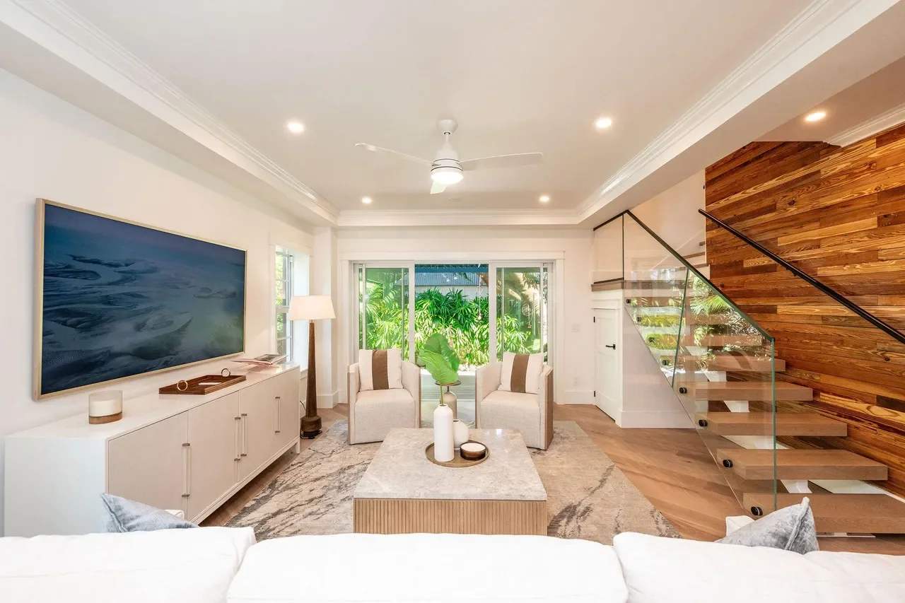 Bright Key West living room with modern coastal furnishings, light wood floors, curated coastal artwork, glass-railed staircase, and sliding doors overlooking lush tropical landscaping.